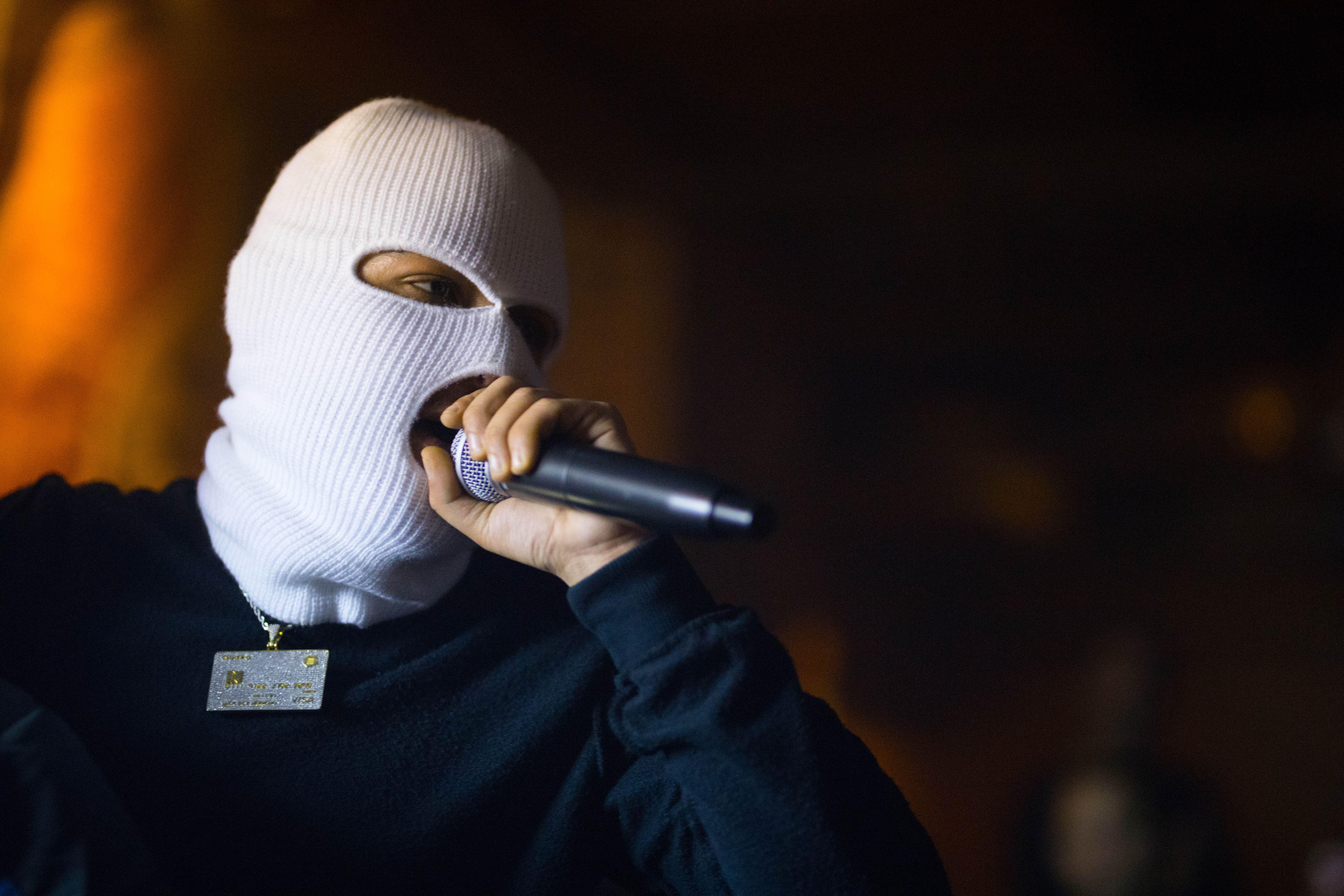 Masked artist wearing a white balaclava performing live on stage with a microphone.