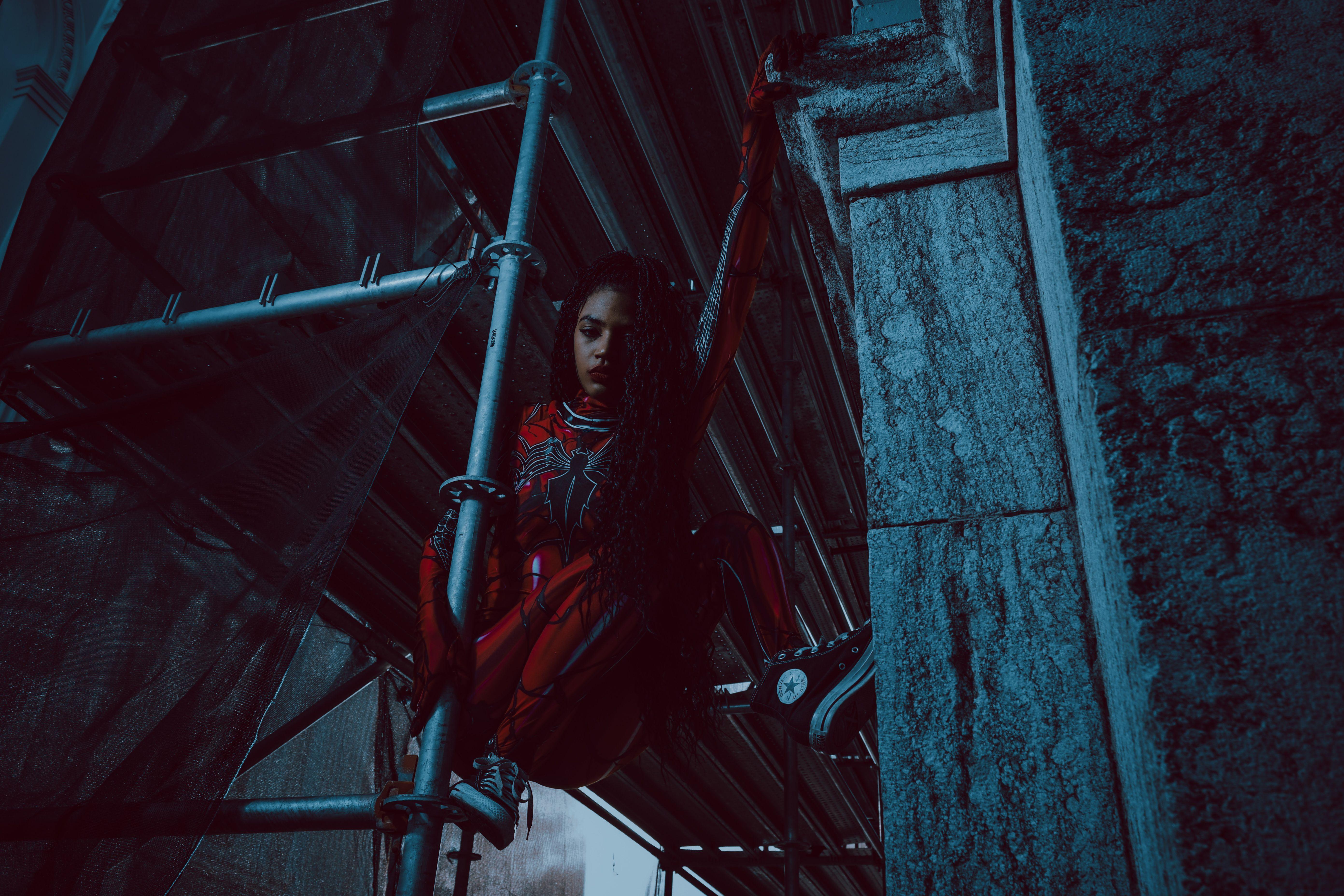 A woman in a Spider-inspired costume clings to scaffolding, showcasing strength and agility.