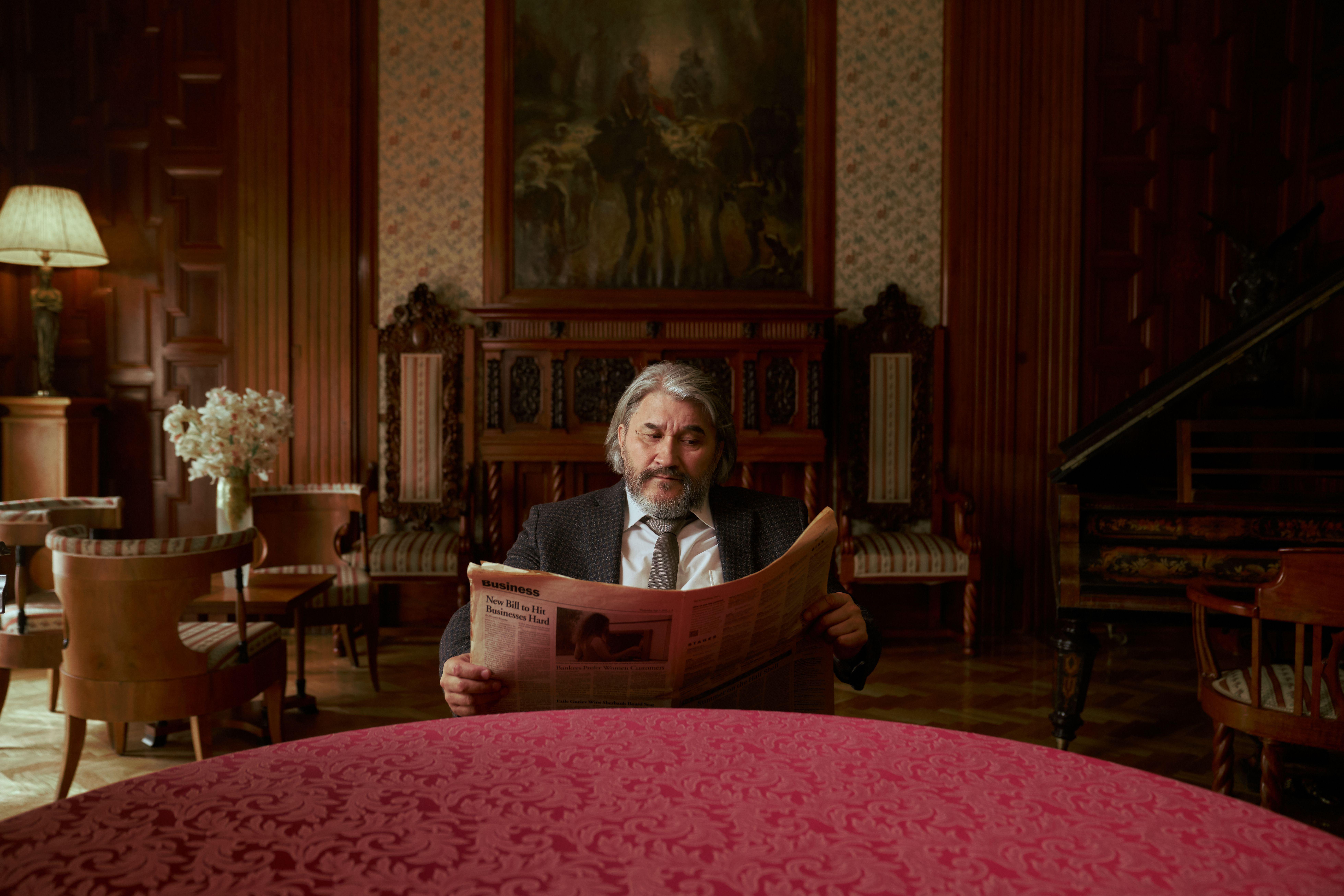 Elderly man reading a newspaper in an opulent, vintage room with classic decor.