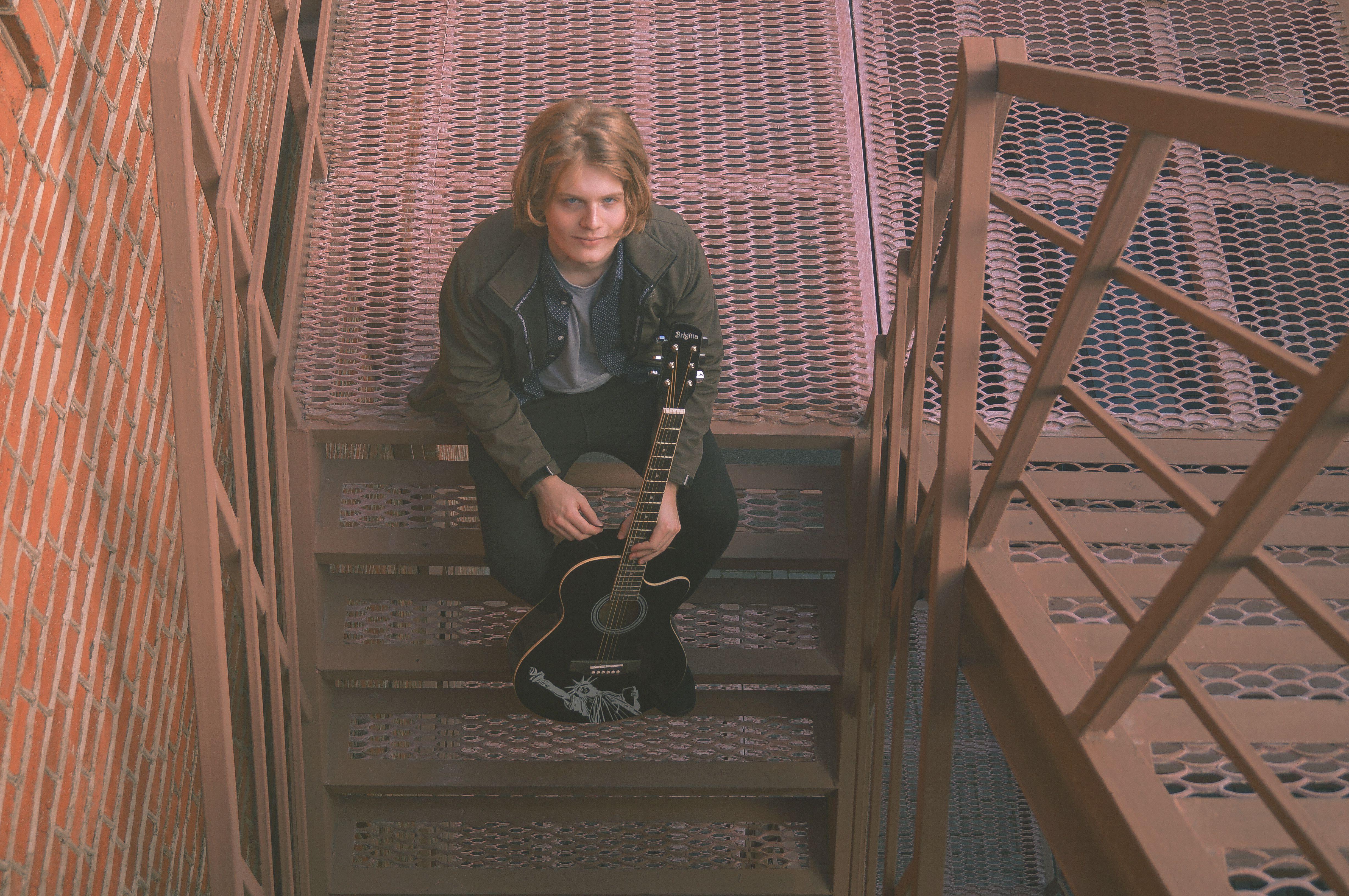 A young man with a guitar sitting on a metal staircase, looking up.