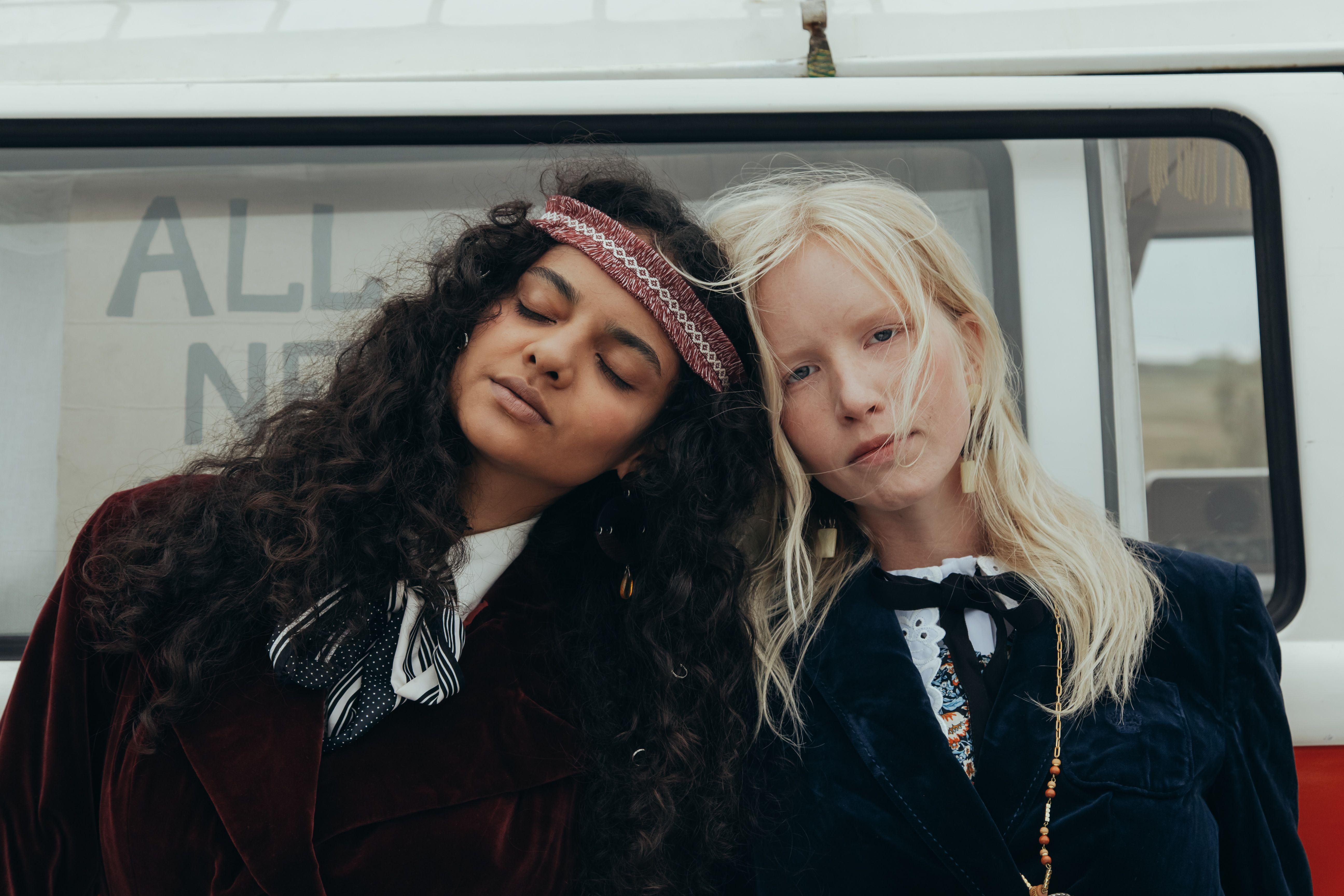 Two stylish women in hippie attire lean against a vintage van, exuding a 60s vibe.