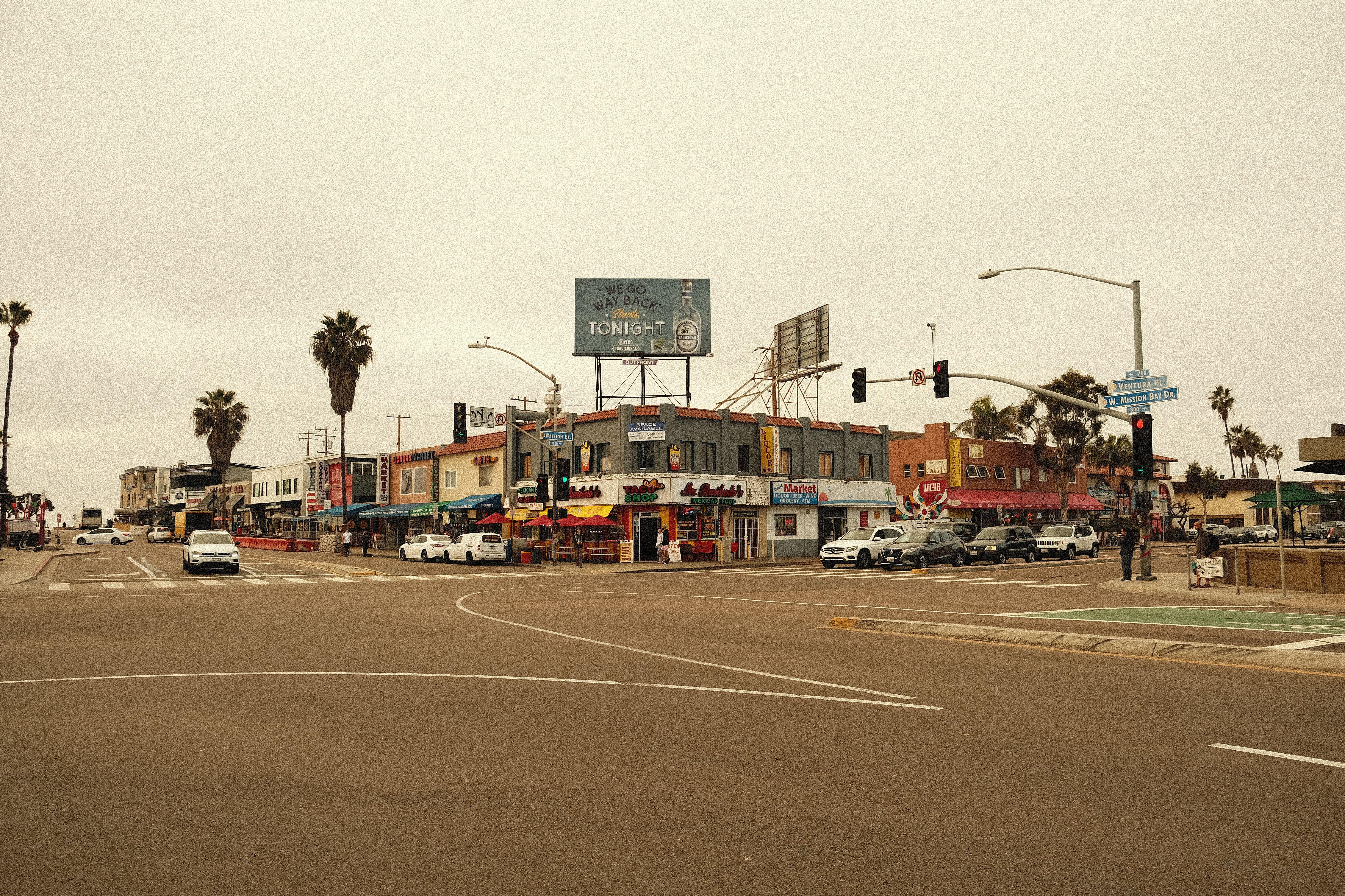 Vibrant city intersection featuring palm trees, shops, and cars under an overcast sky.