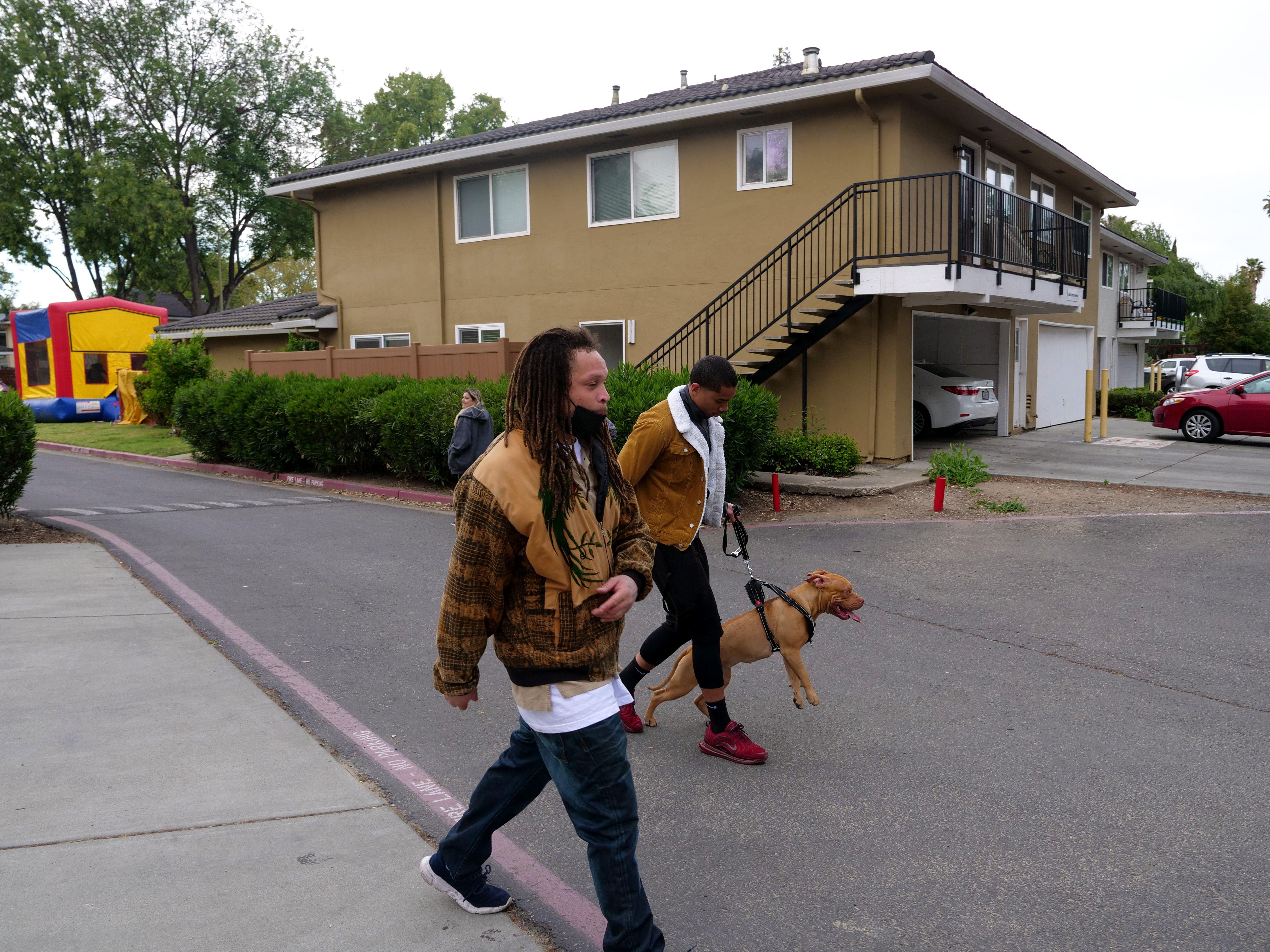 Two people walk a dog in a suburban neighborhood with residential buildings.
