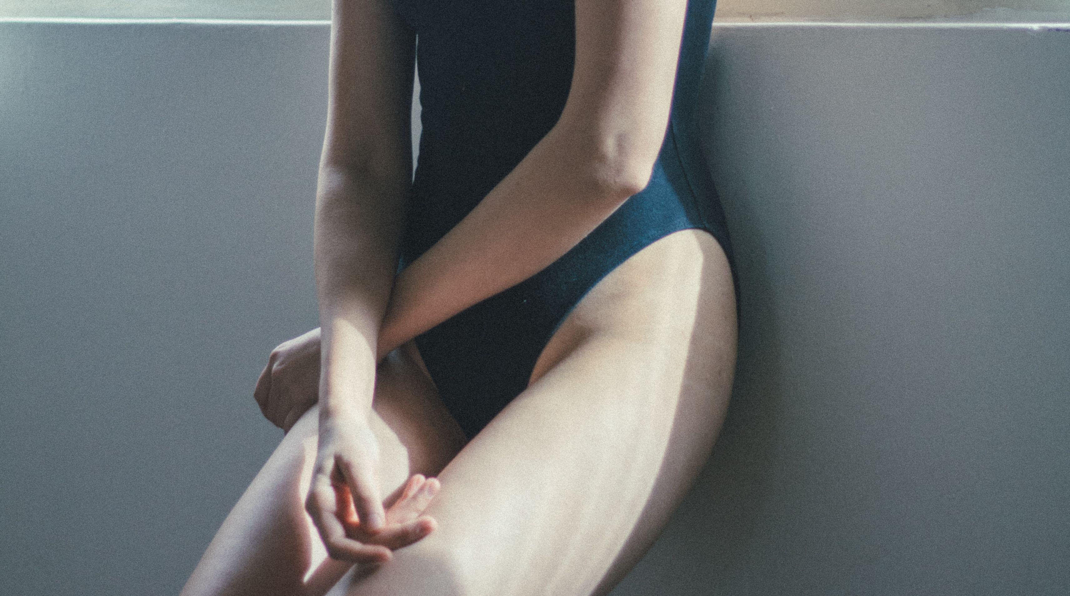 Close-up of a woman in a black swimsuit leaning against a gray wall with soft lighting.