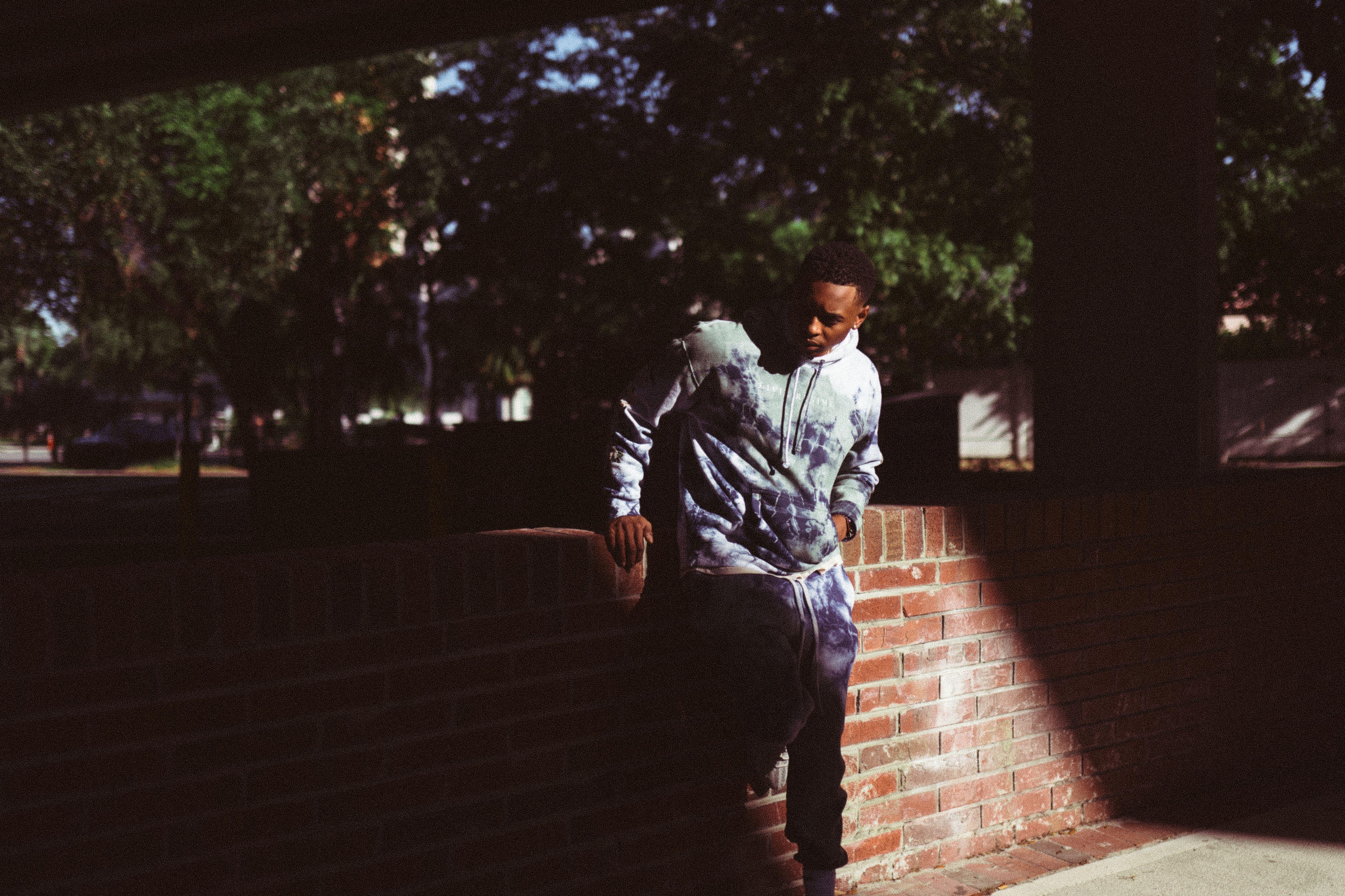A young man in a casual sweatshirt leans against a brick wall in a sunlit park.