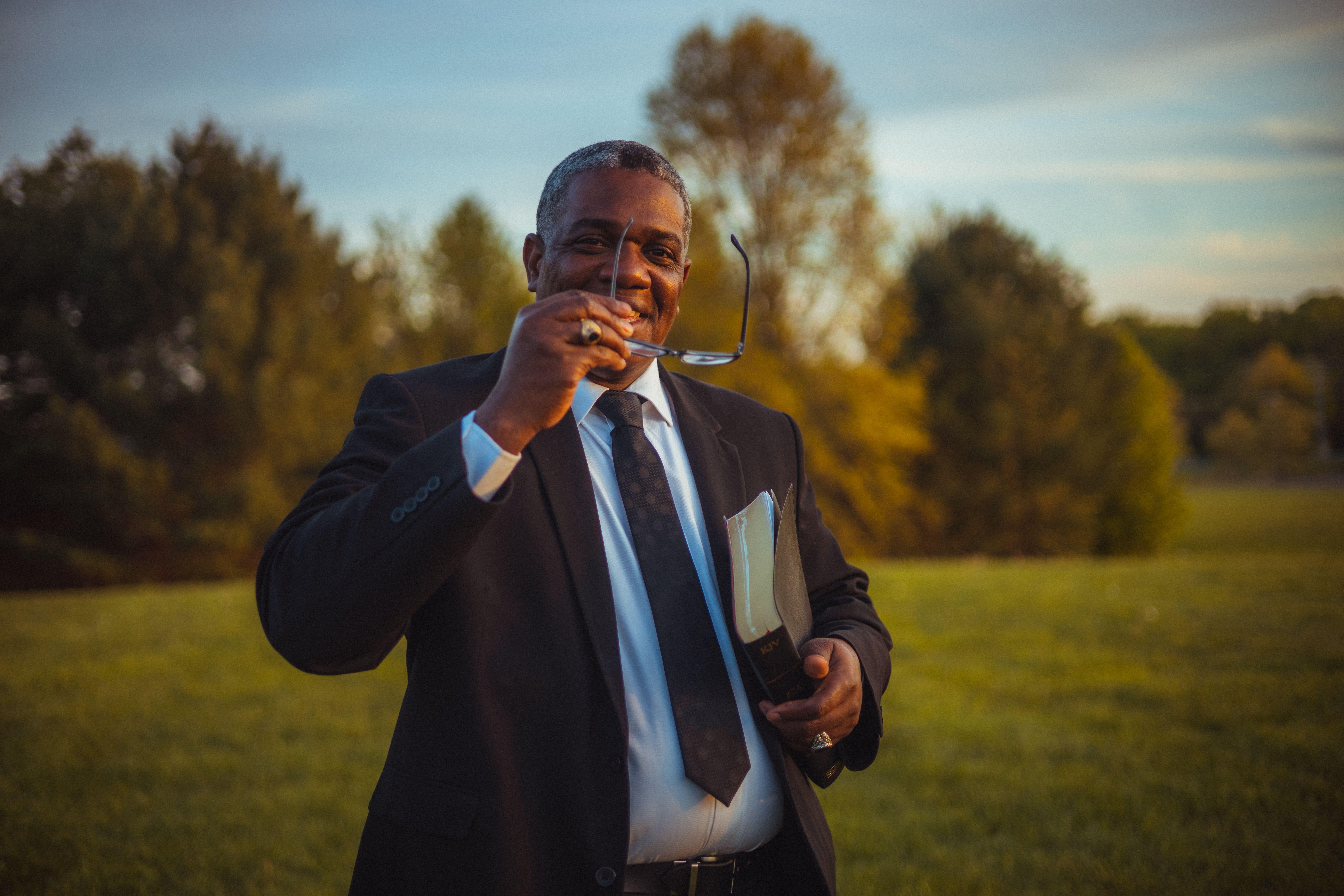 African American man in a suit smiling outside with eyeglasses and book.