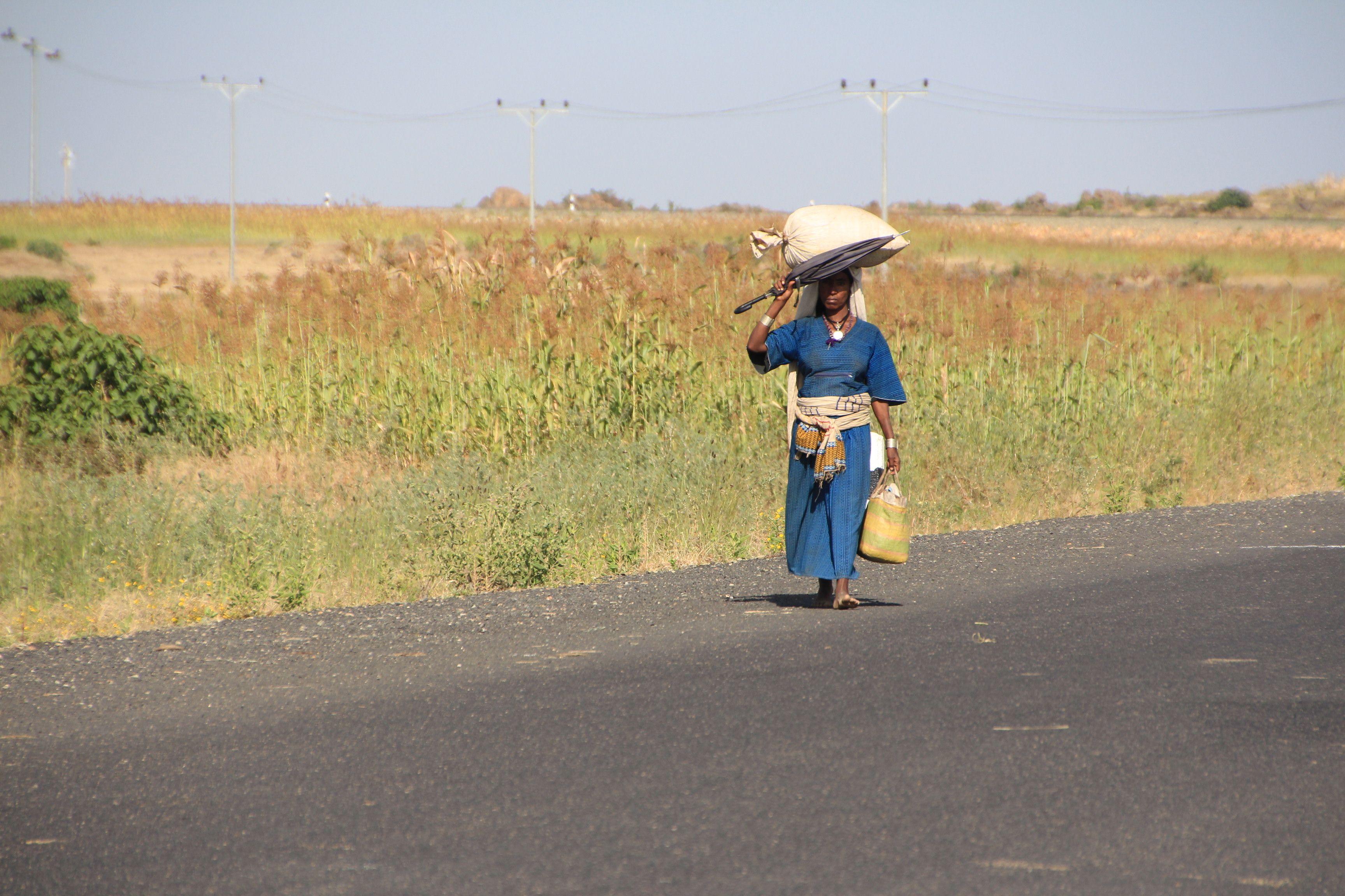 A barefooted Ethiopian woman walks along a rural road carrying goods on her head.