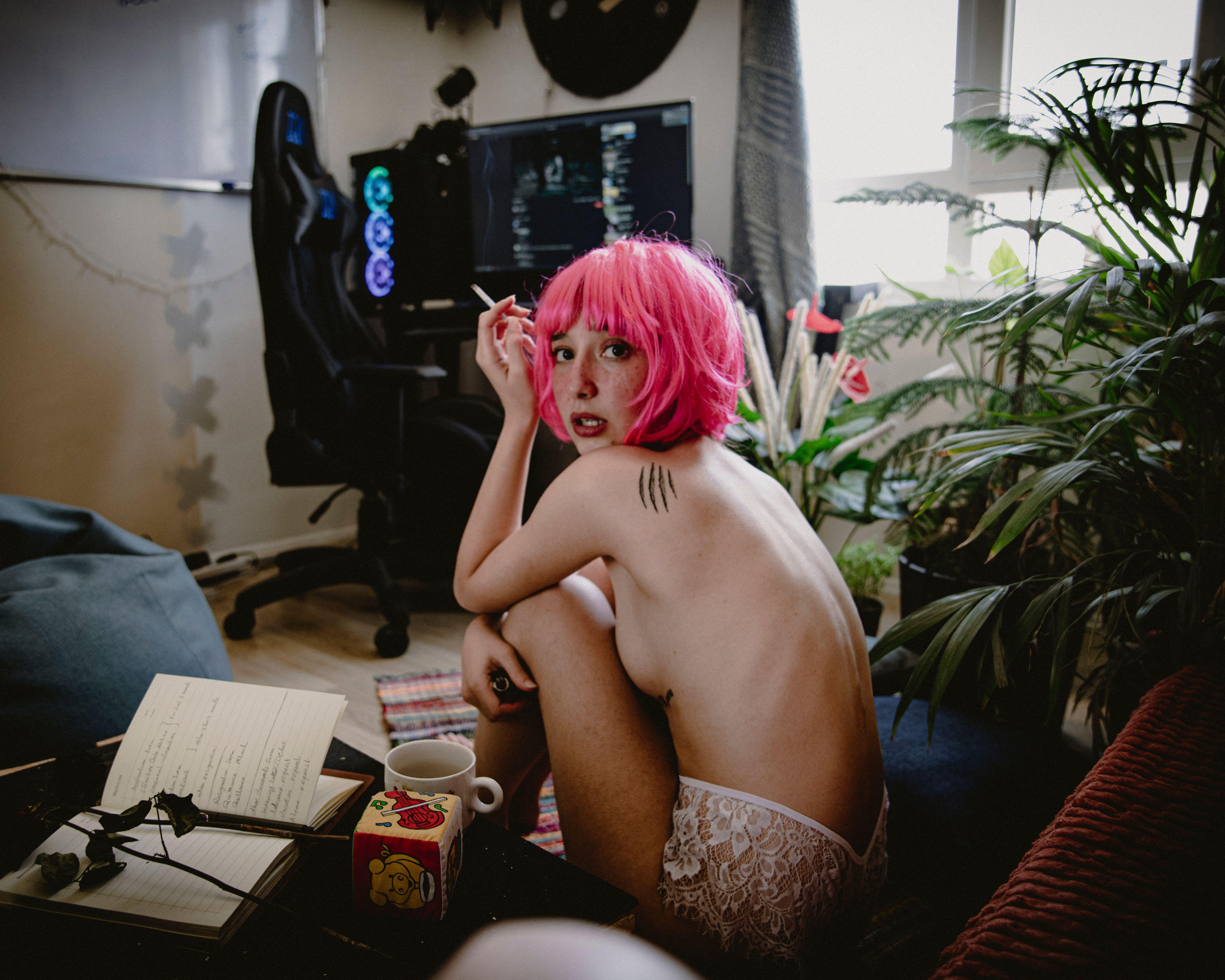 A young woman with pink hair sits indoors, surrounded by plants and notebooks, creating a cozy atmosphere.