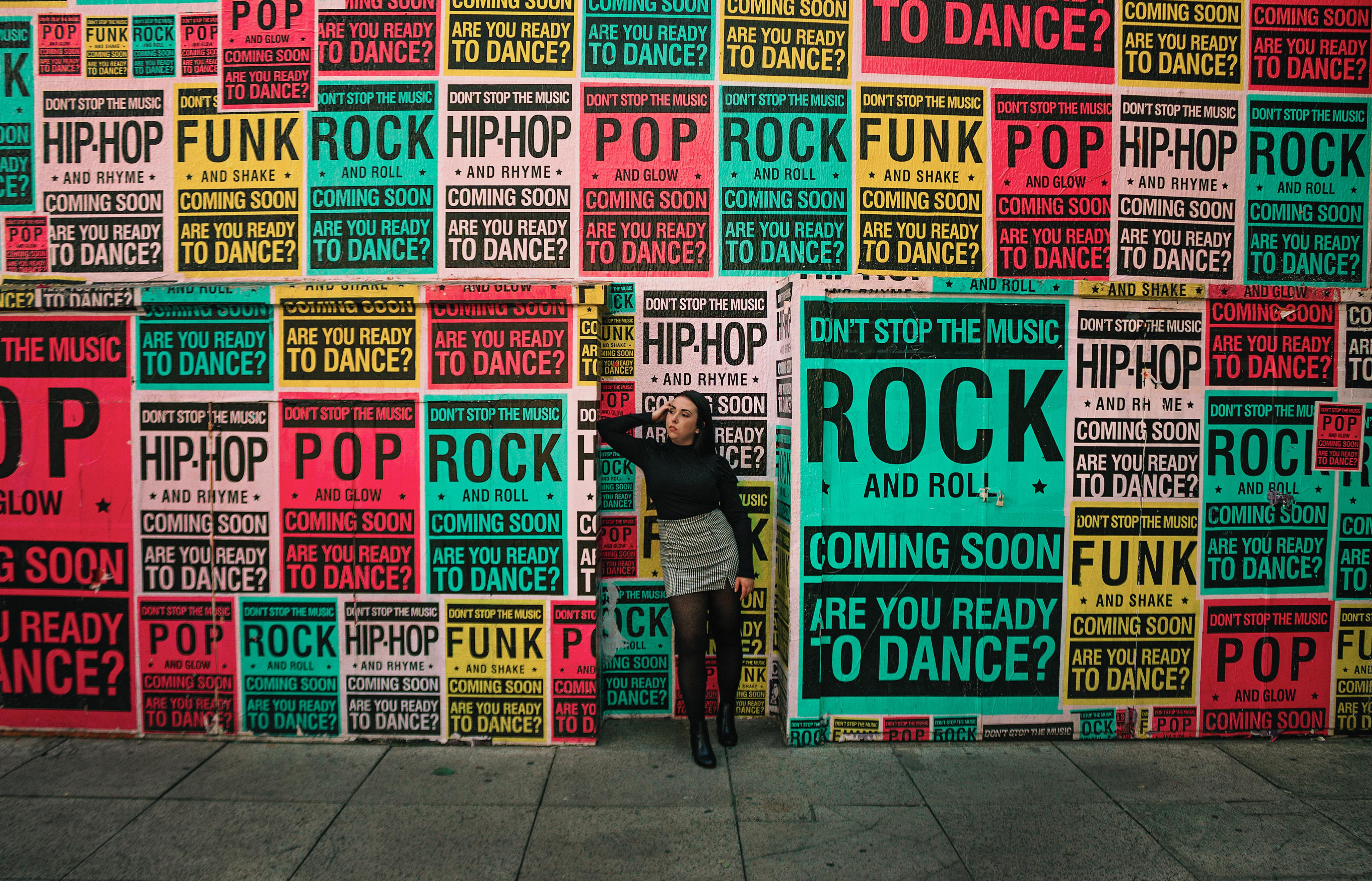 Woman leans against a wall covered in colorful music genre posters. Urban scene.