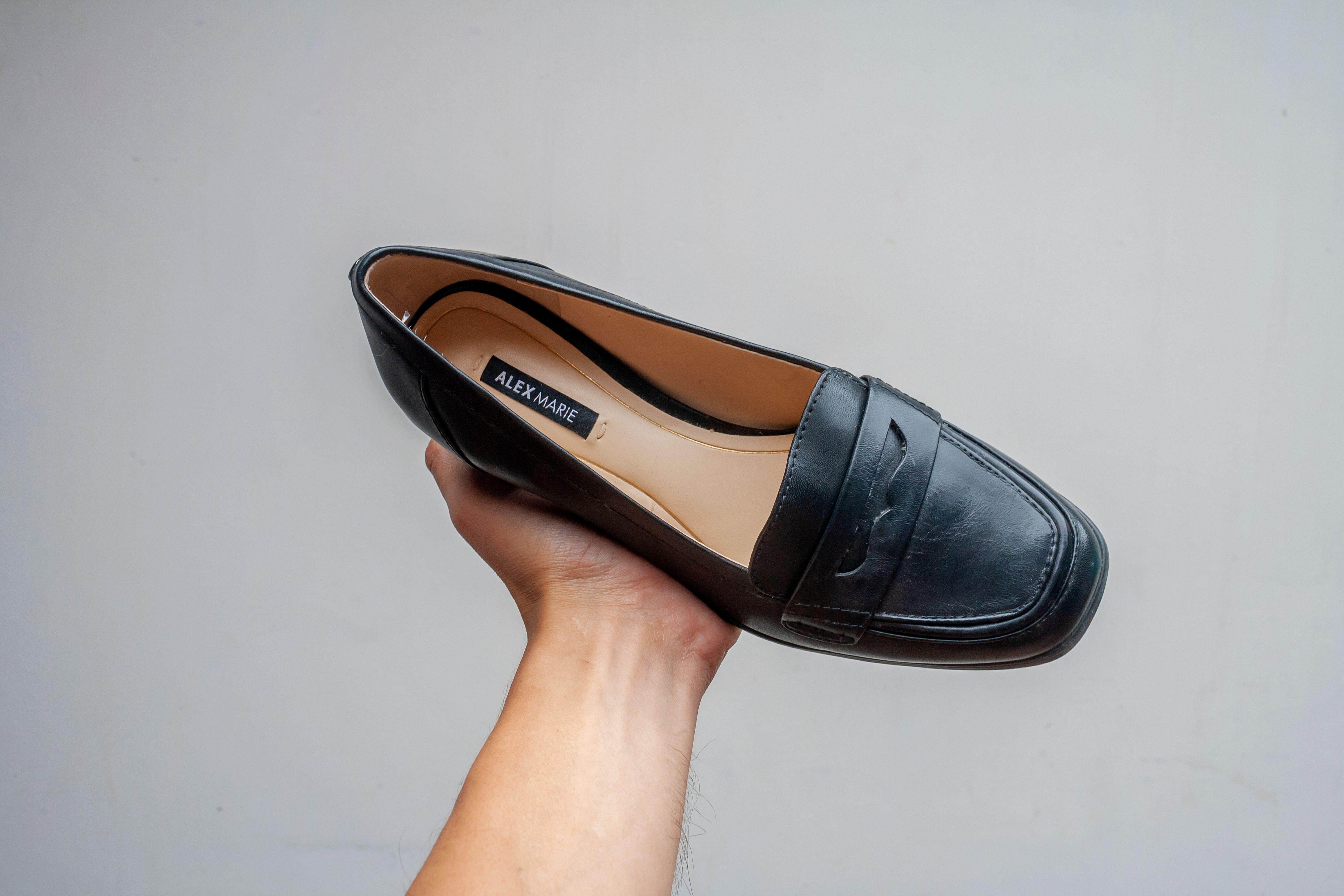 Close-up of a hand holding a black leather shoe by Alex Marie against a plain background.
