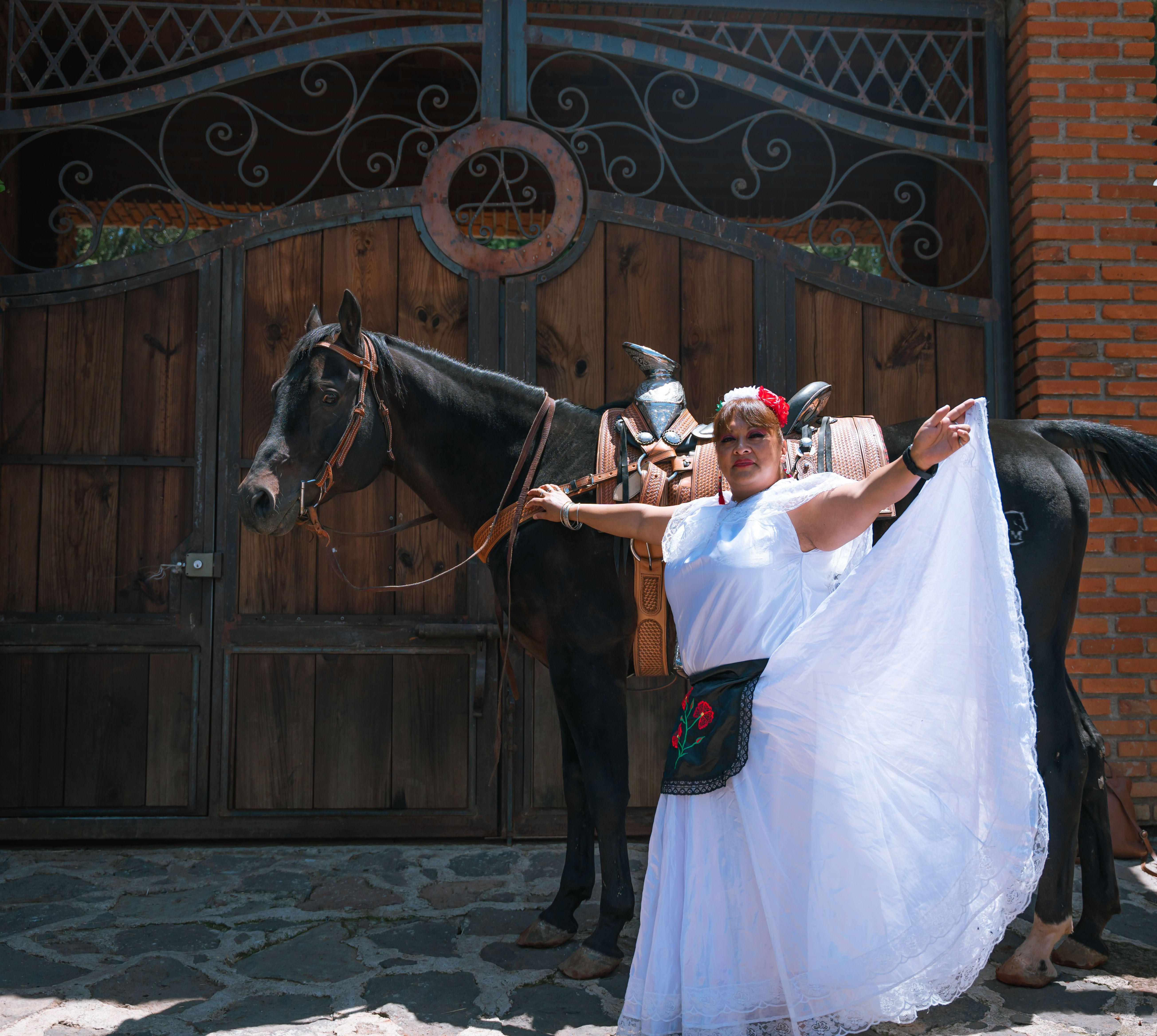 A woman in a white dress and traditional attire poses joyfully beside a black horse.
