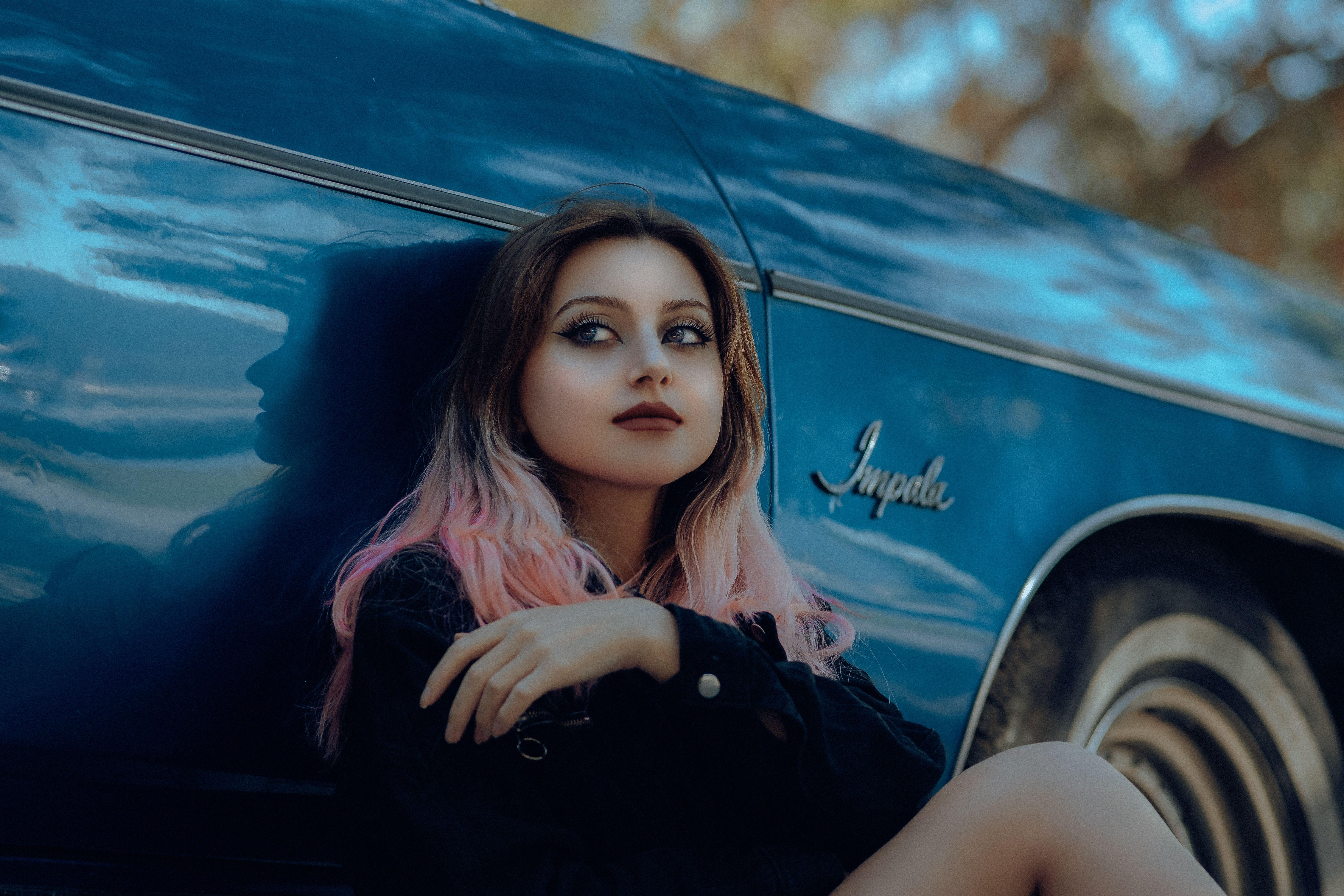 A young woman with pink hair leans against a vintage blue Impala car, with a thoughtful expression.