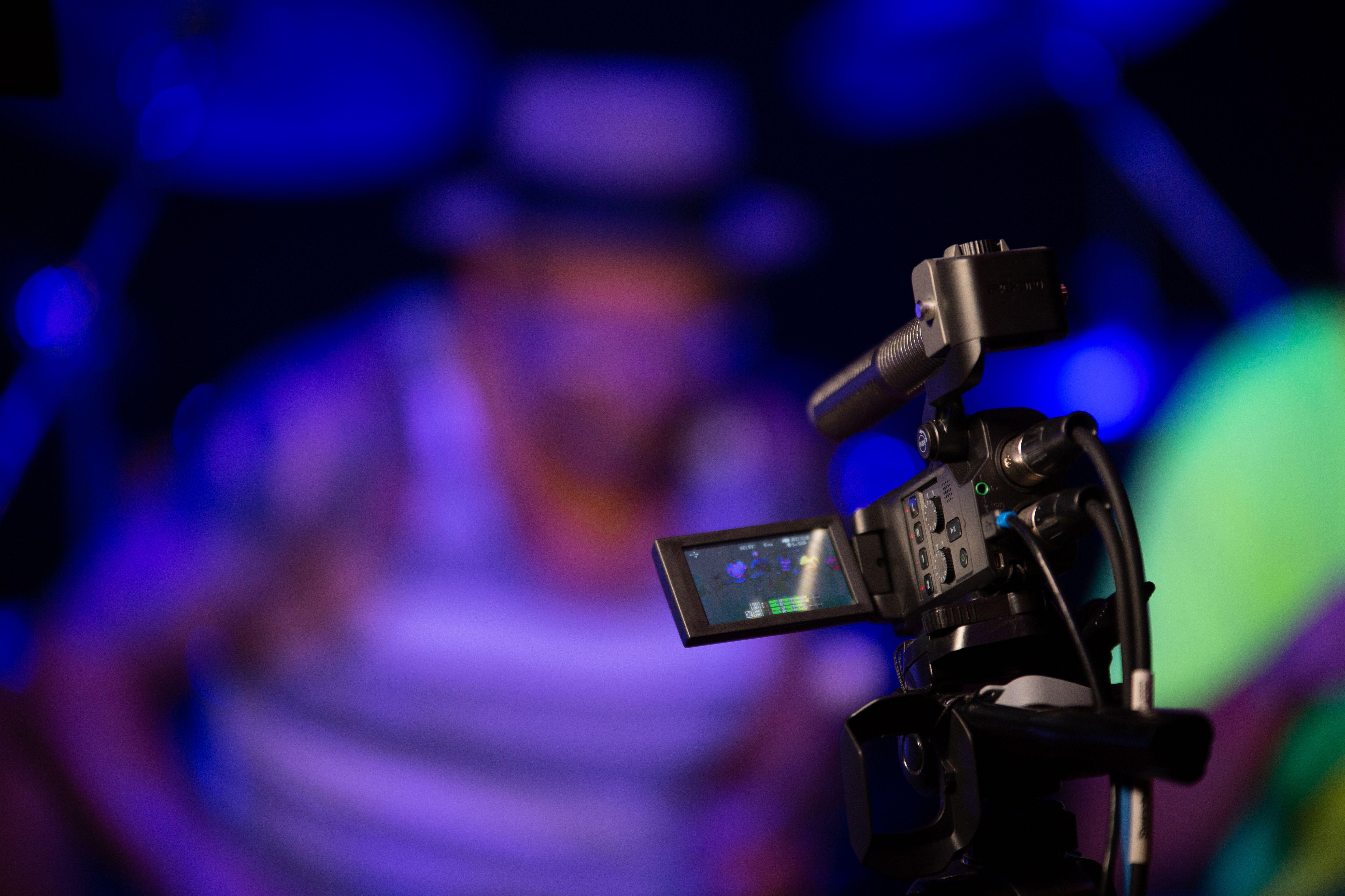 Close-up of a video camera capturing a live event with blurred background, illuminated by vibrant lights.