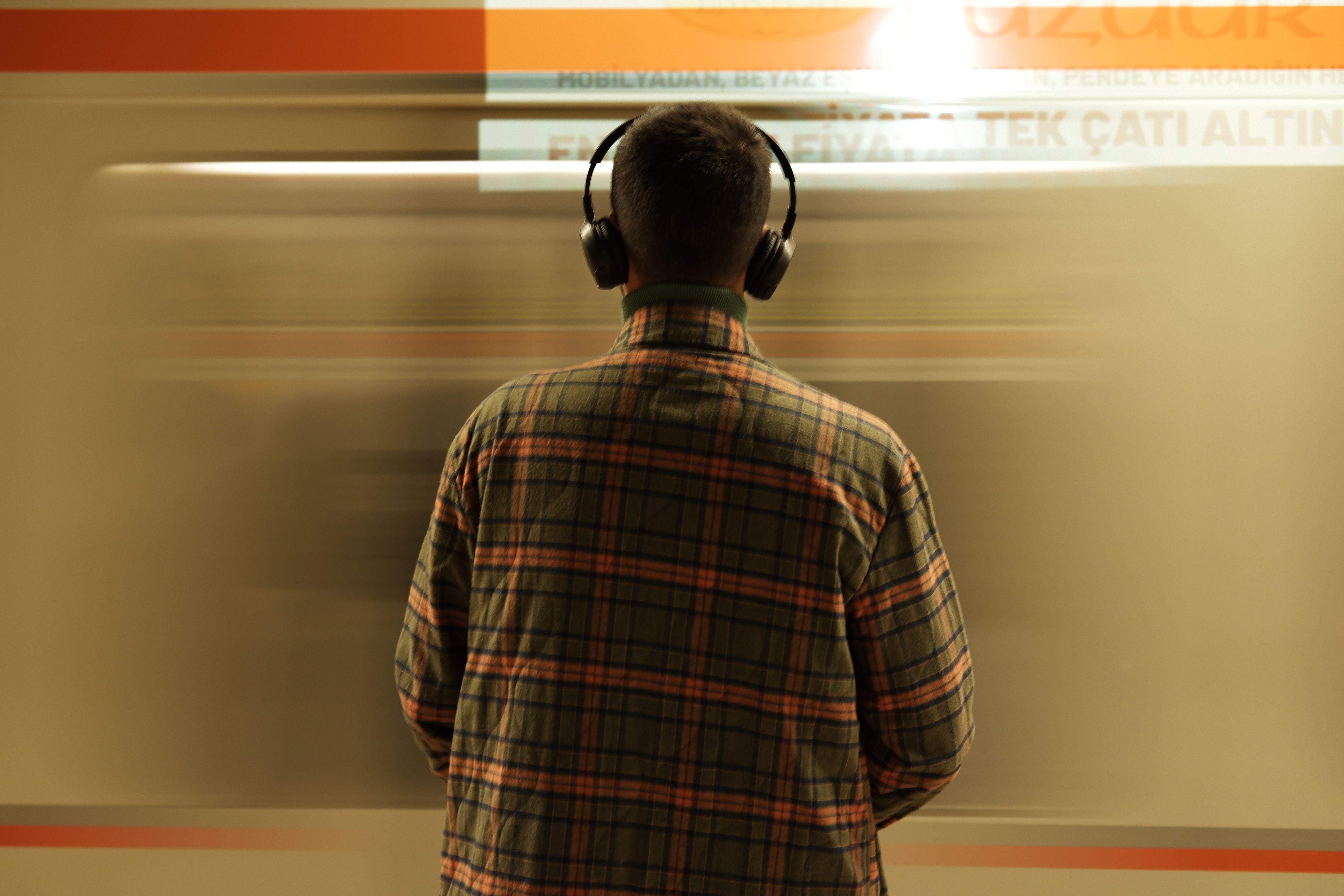 A man wearing headphones stands at a subway station, observing a passing train.