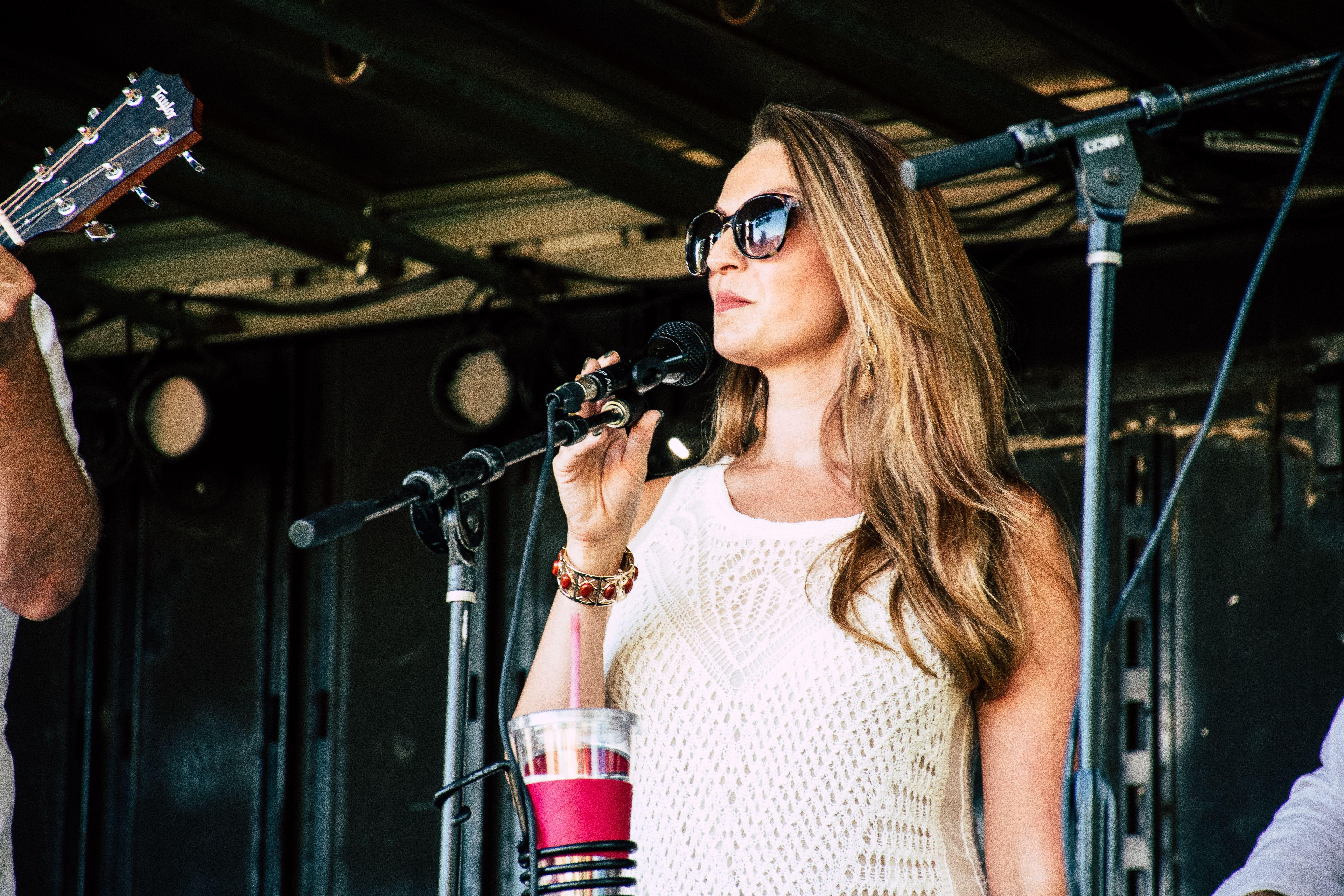 A woman in sunglasses sings on stage during an outdoor concert, exuding confidence and style.