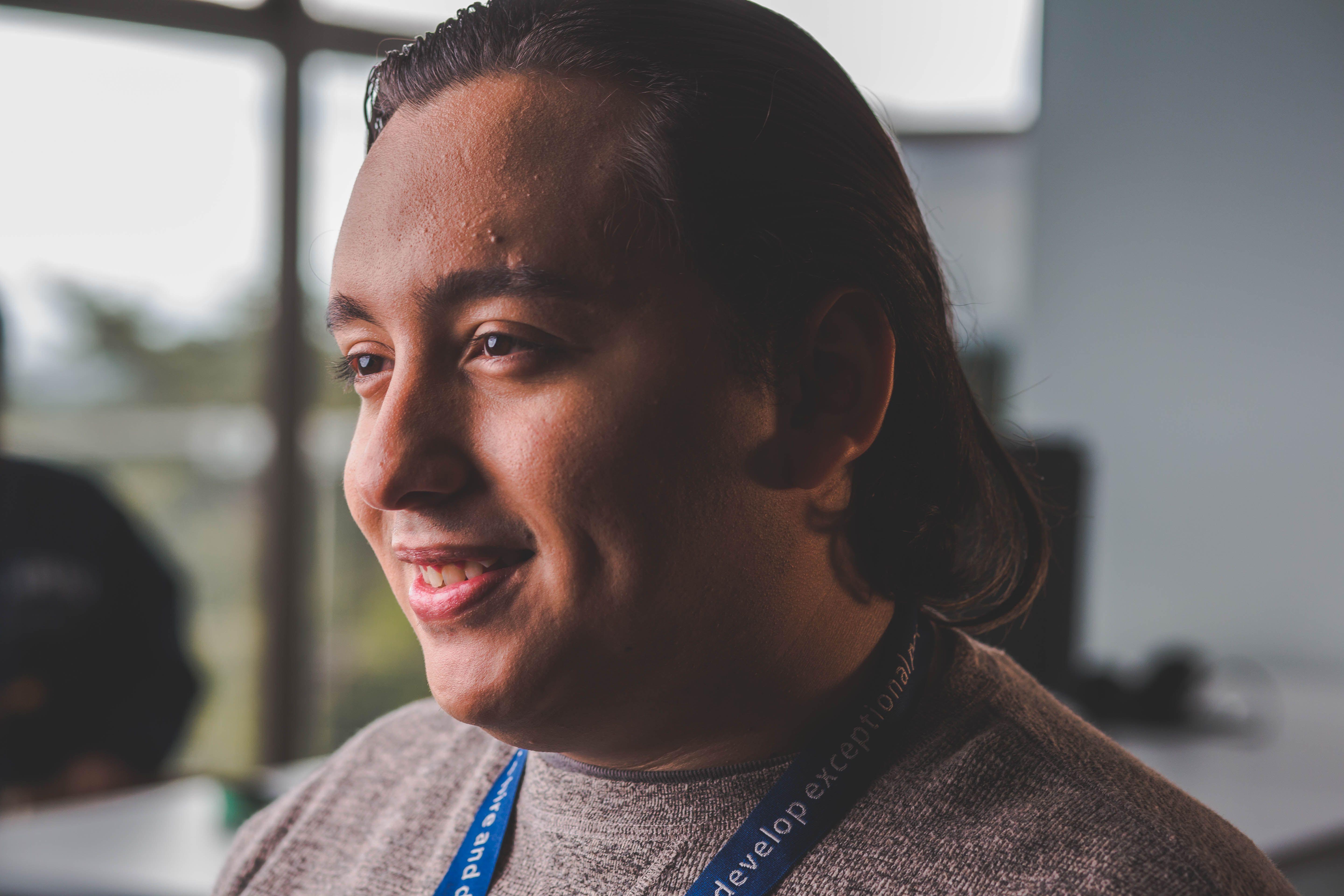 Portrait of a smiling man with long hair, wearing a sweater, in a bright office setting.