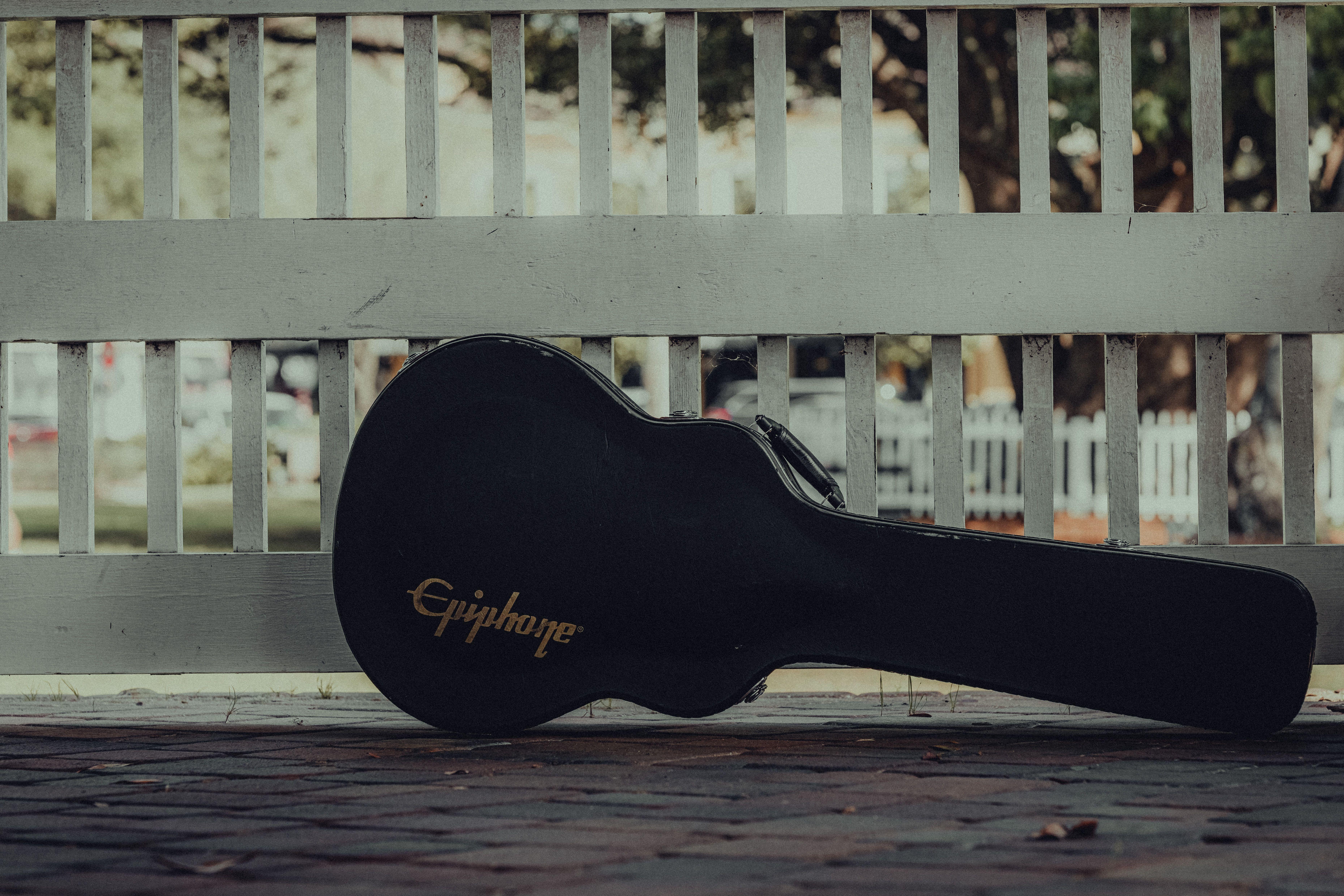 A guitar case rests against a white fence on a pavement outdoors.