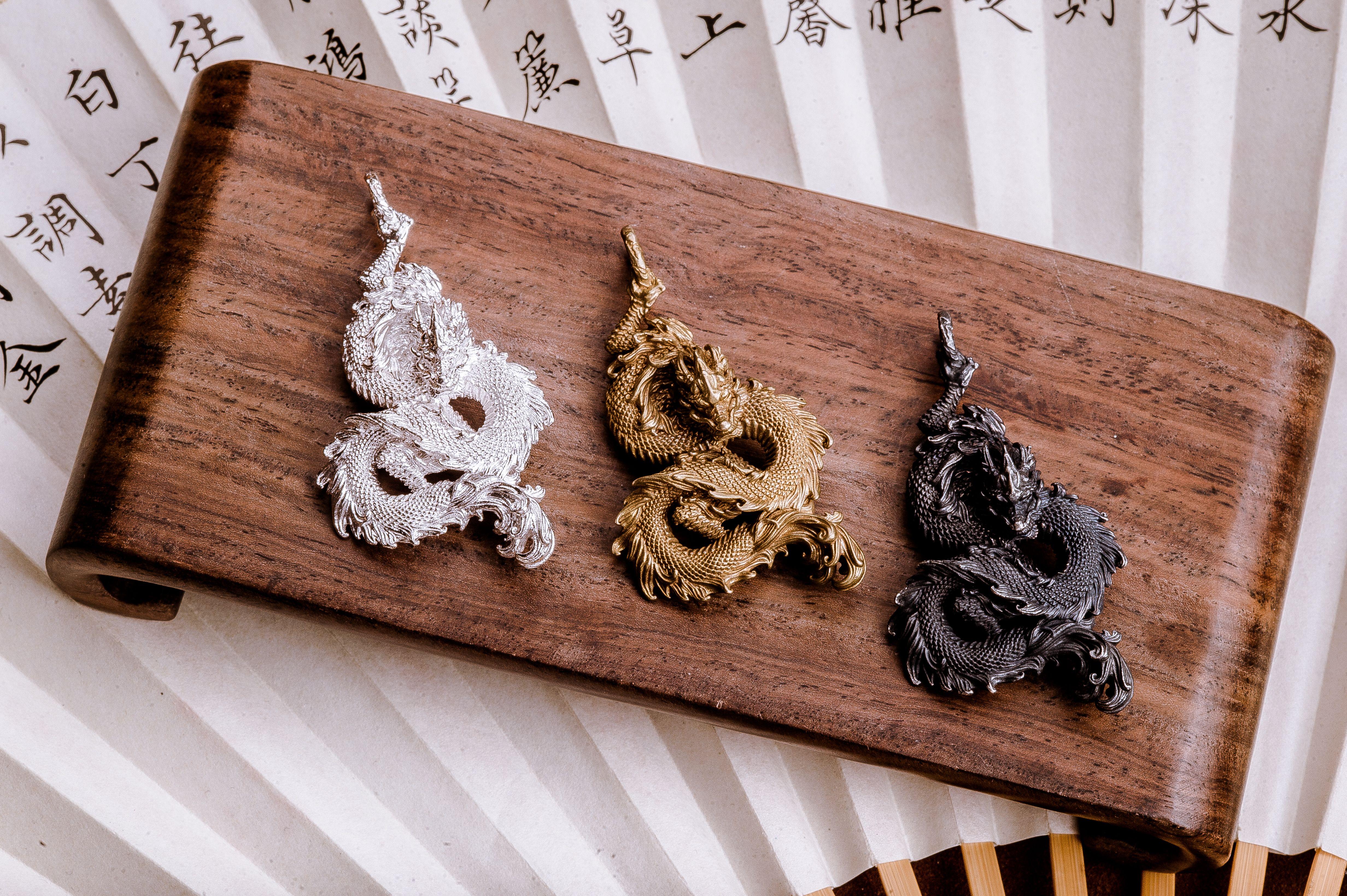 Three intricately designed dragon pendants in silver, gold, and black on a wooden stand.