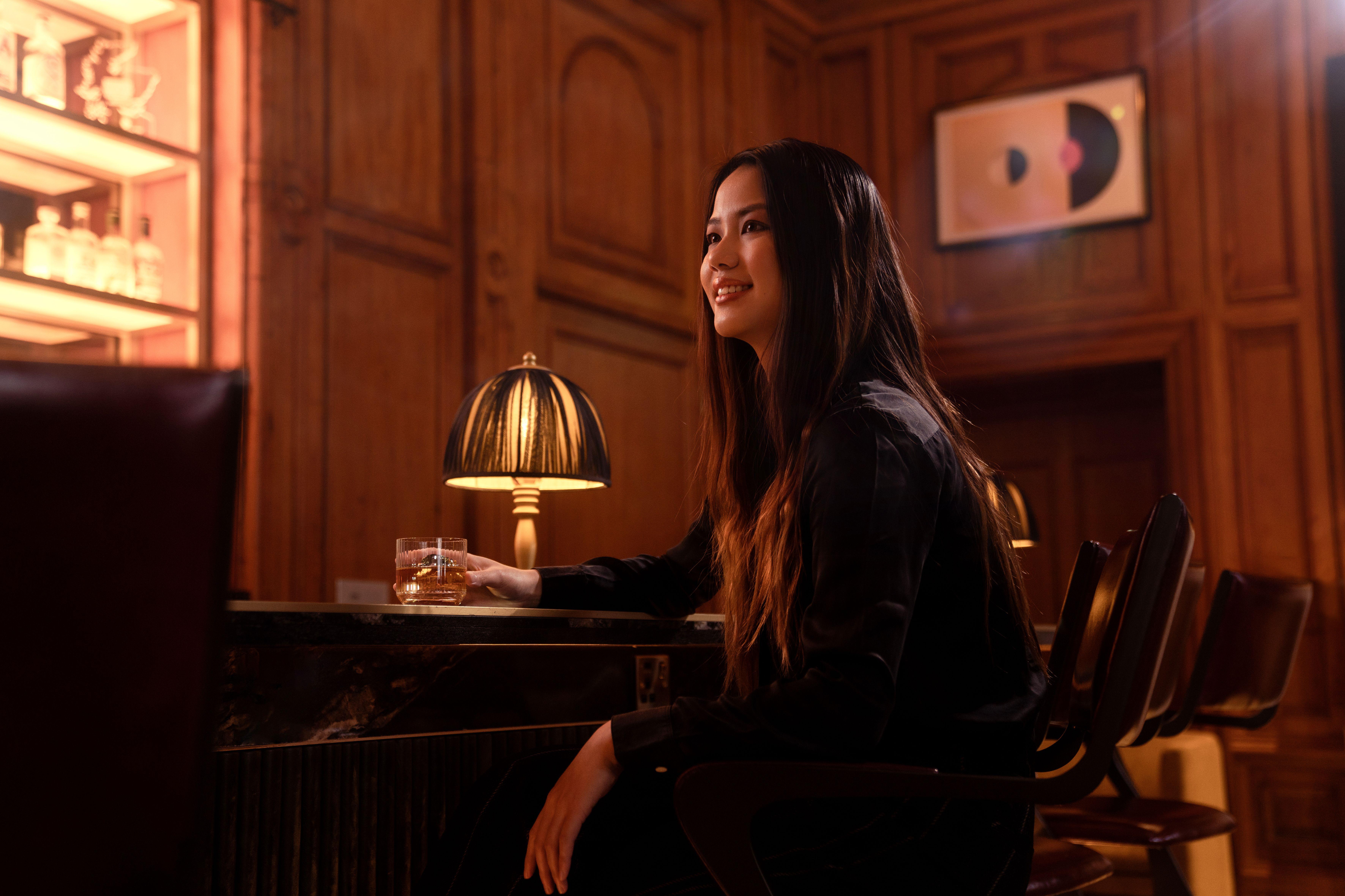 Woman with long hair sipping whiskey at a stylish bar in warm lighting, capturing elegance and relaxation.