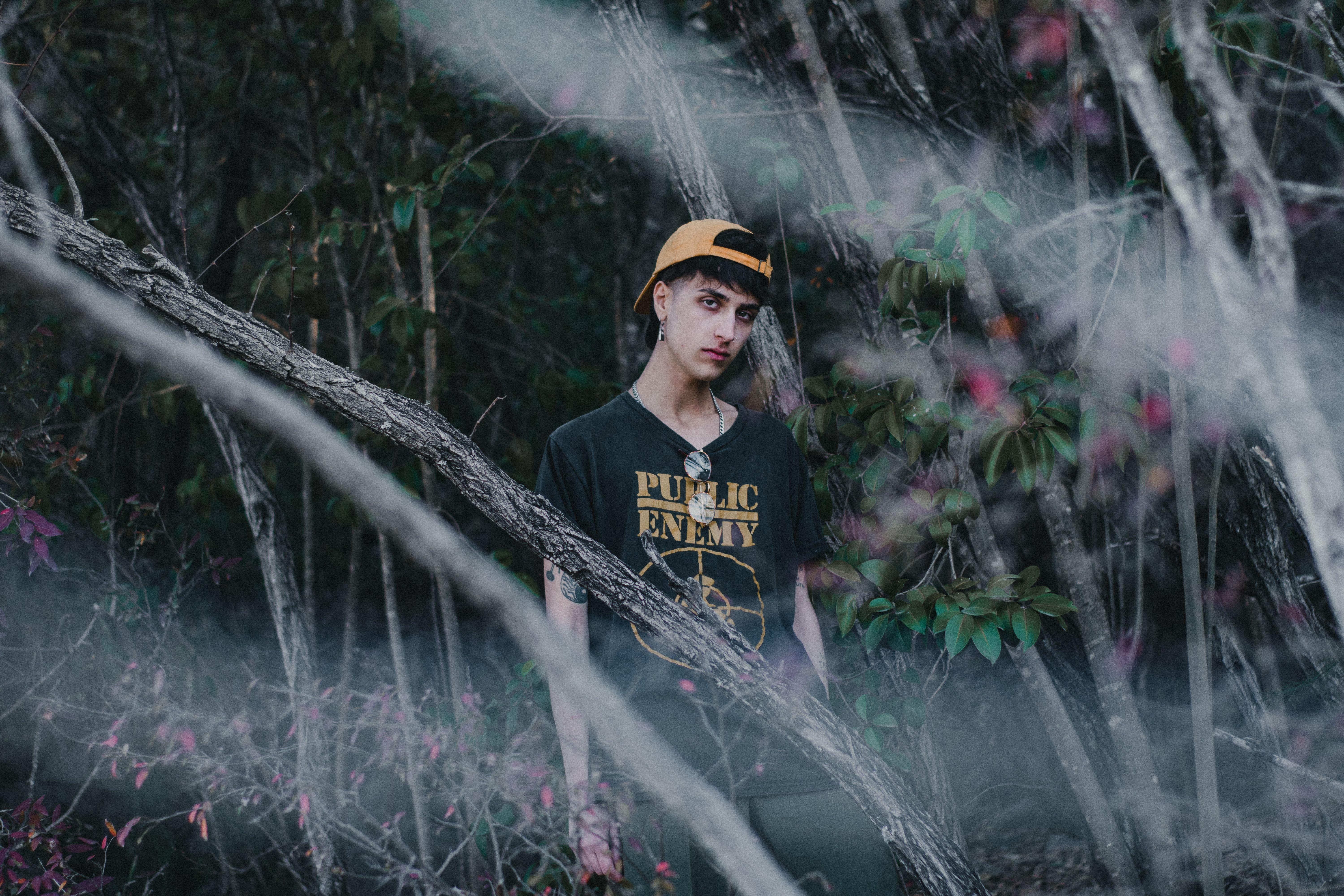 Man in black t-shirt and cap poses amid forest trees. Moody artistic portrait.