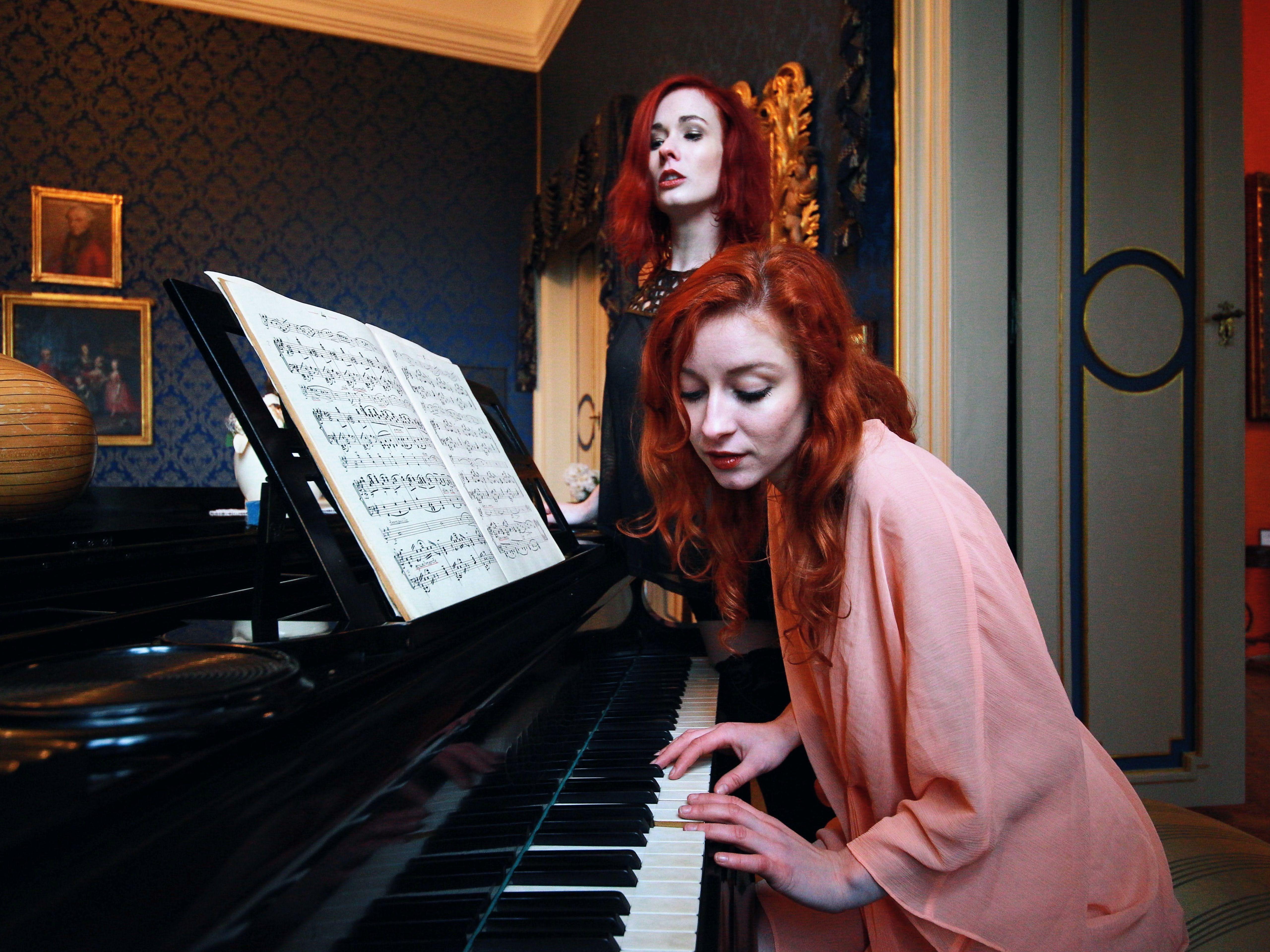 Red-haired women engage in a piano duet within an elegant, ornate room setting.