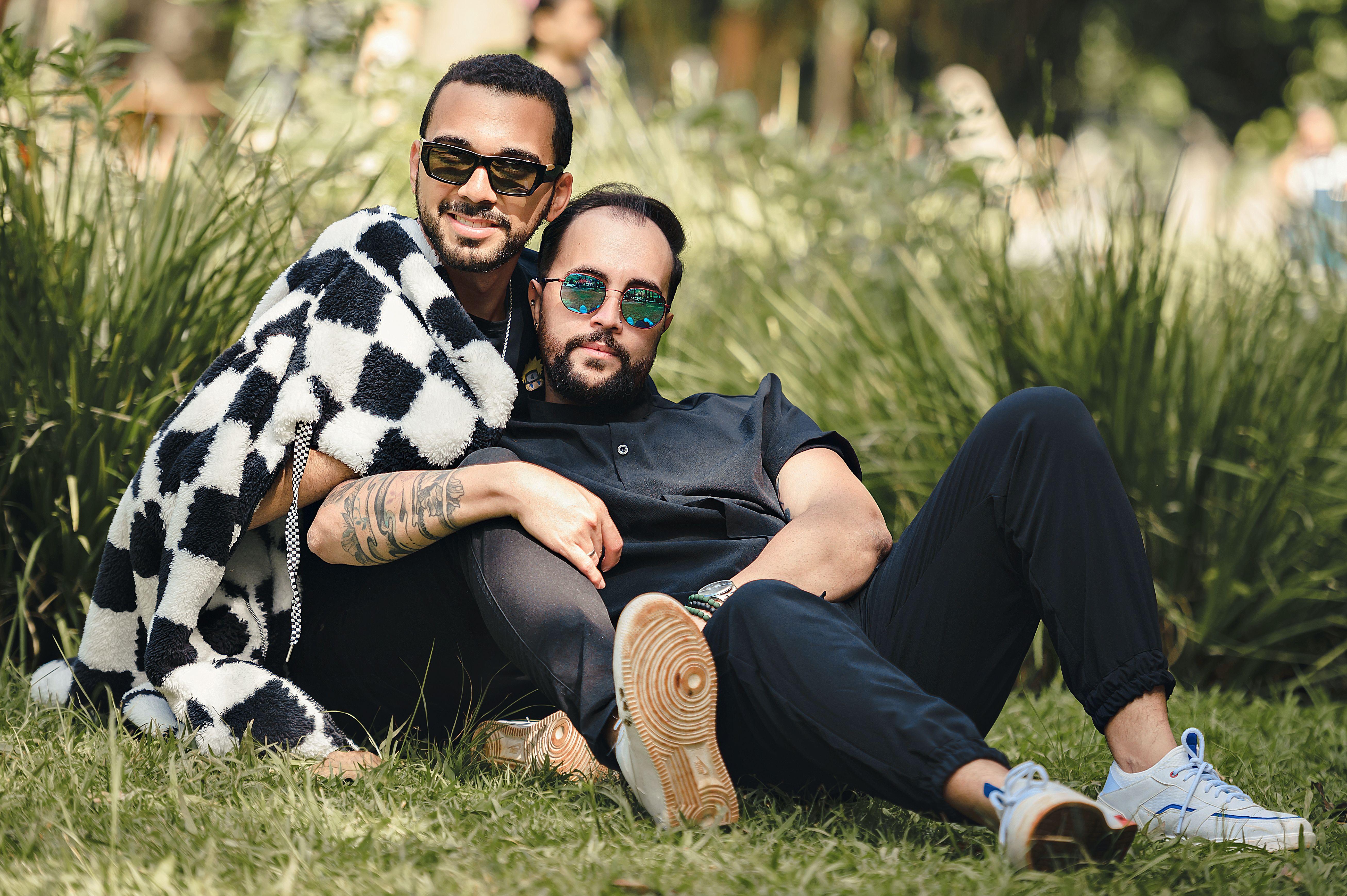 Two men wearing sunglasses, enjoying a relaxed moment on a grassy field with greenery in the background.