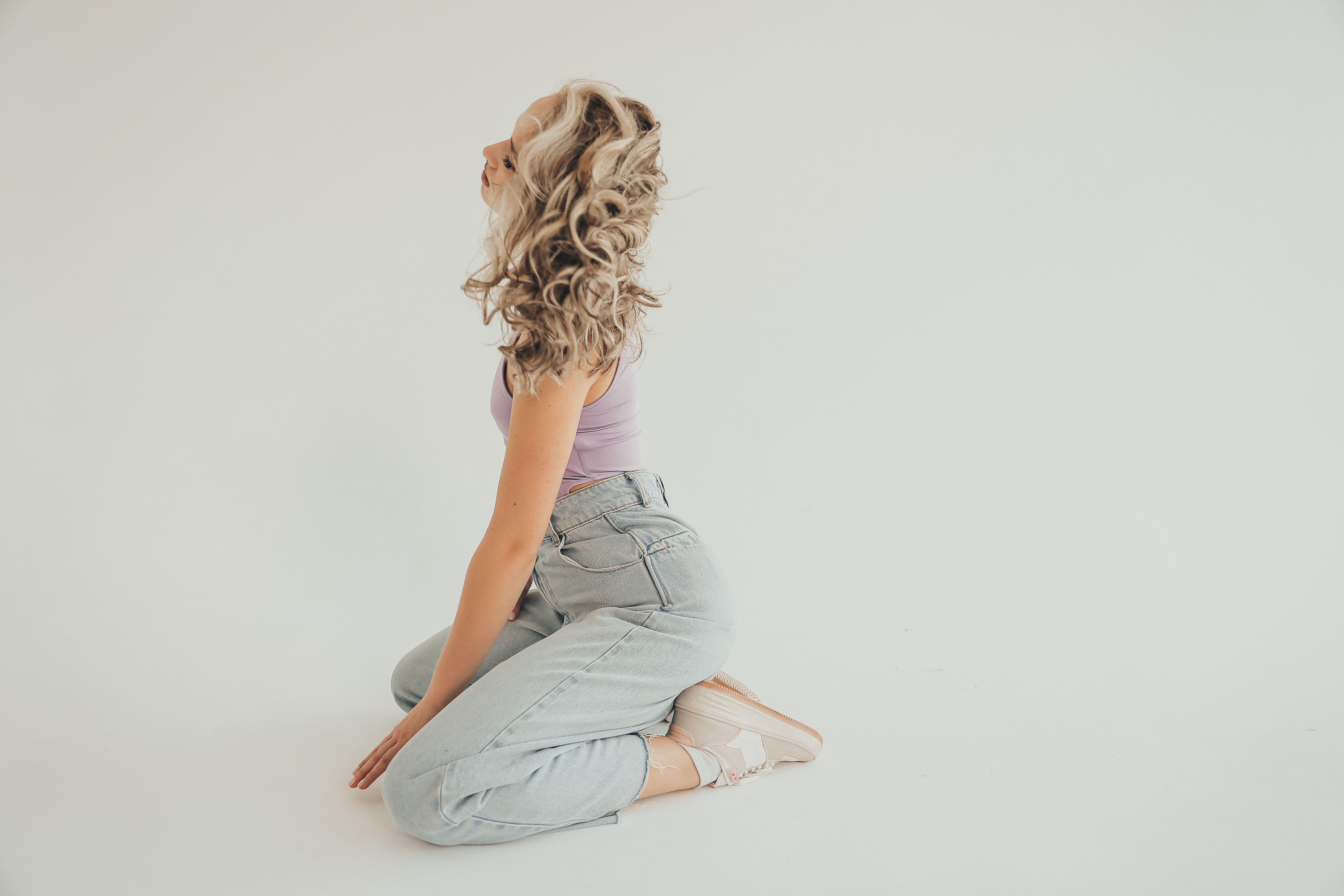 A fashionable woman with curly hair poses in denim jeans against a white background, exuding style and grace.
