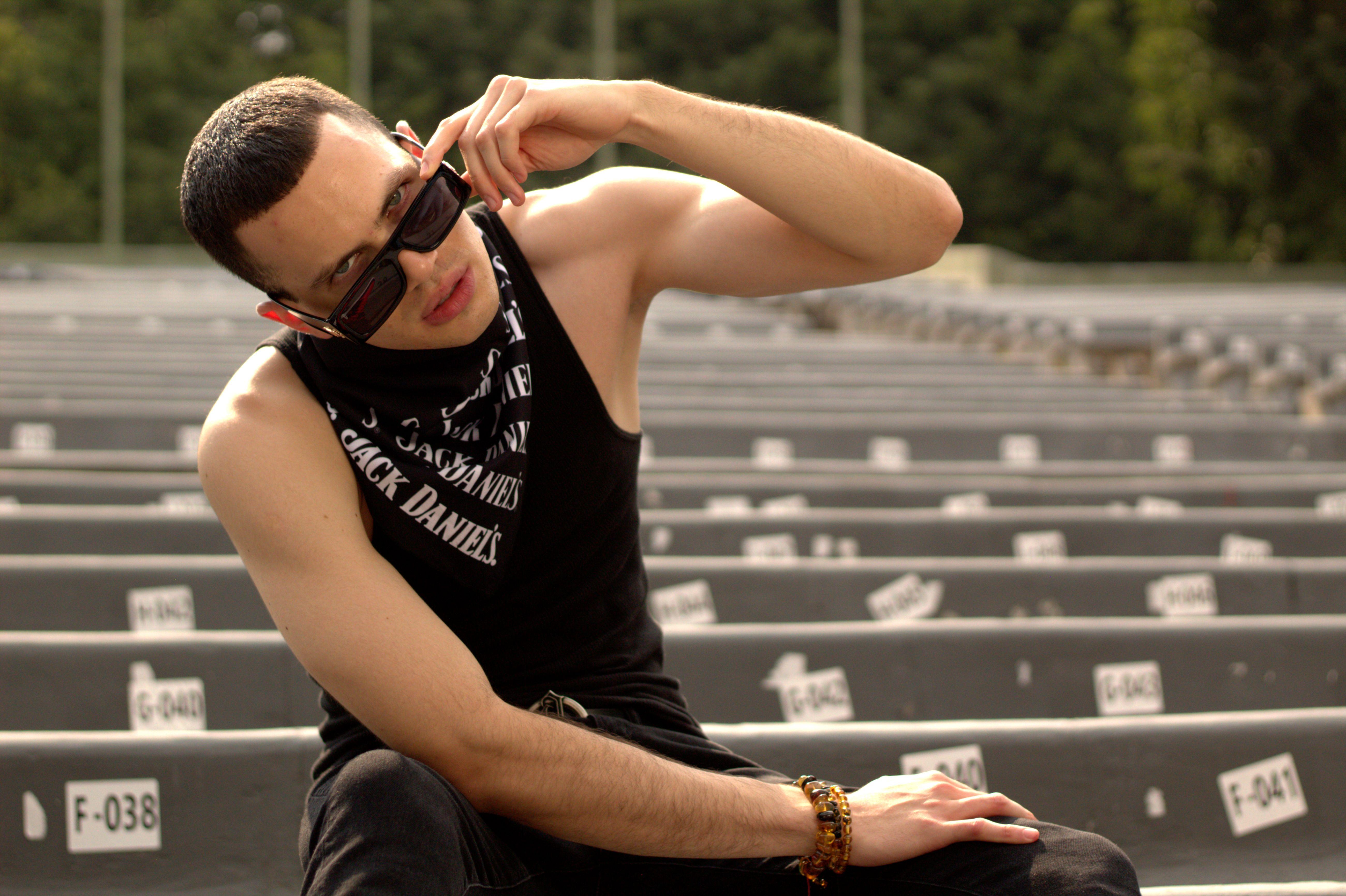 Fashionable young man in black tank top sitting confidently on empty bleachers, adjusting sunglasses.