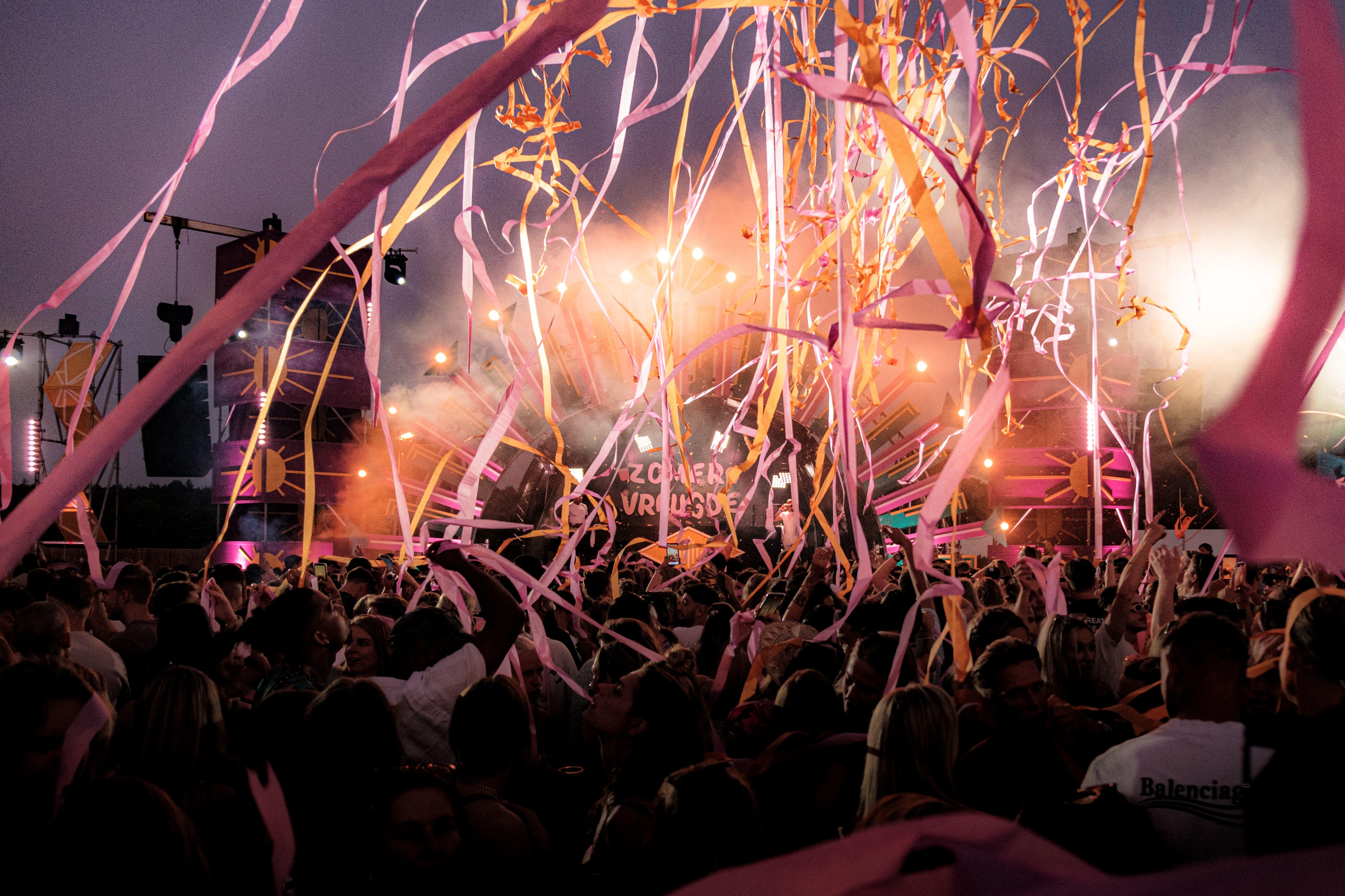 Crowd enjoying a vibrant festival with colorful ribbons and stage lights under a twilight sky.