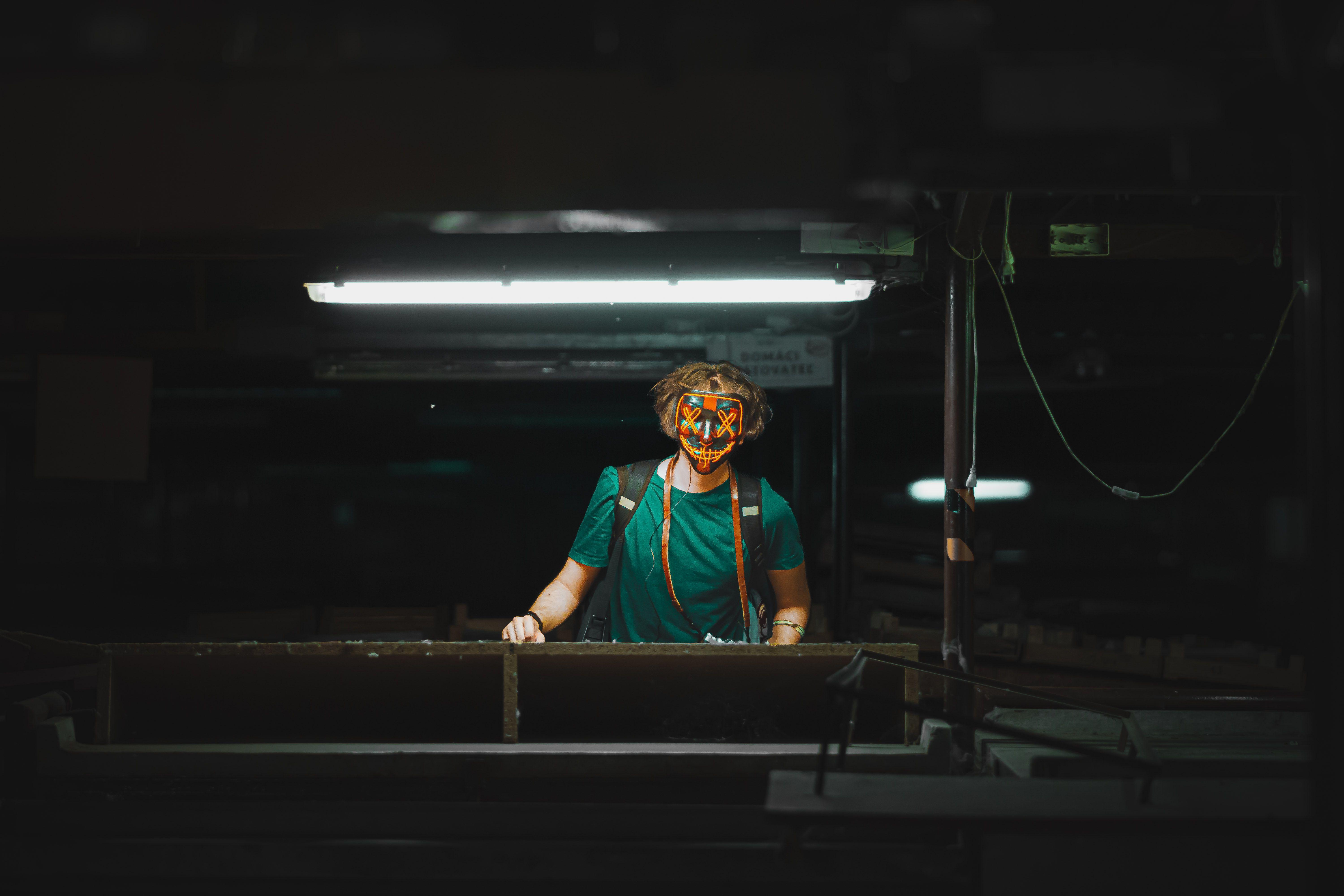 A masked person stands under a fluorescent light in an industrial environment, creating a mysterious and moody atmosphere.