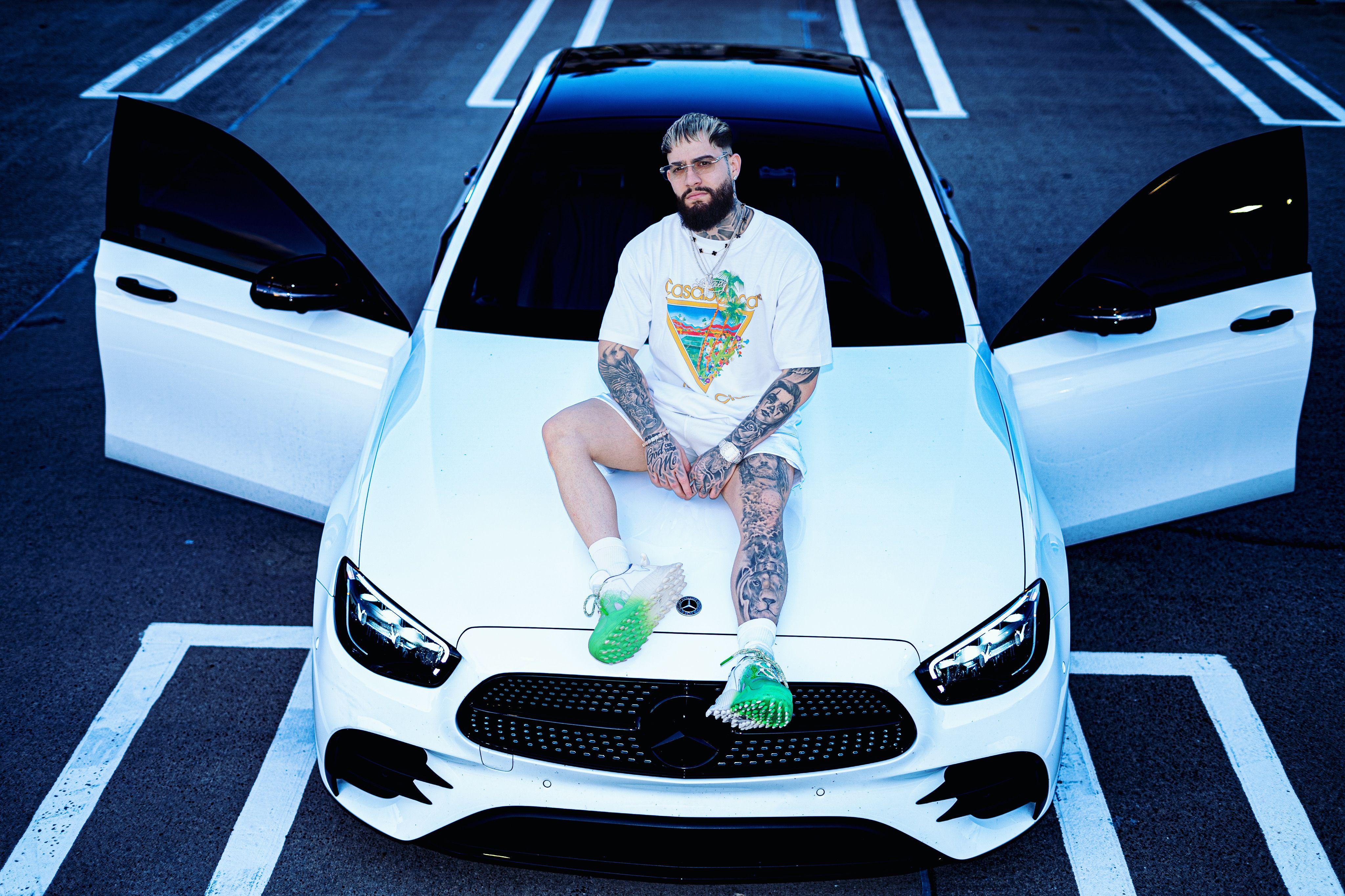 Tattooed man sitting on a white luxury car with open doors in an urban parking lot.