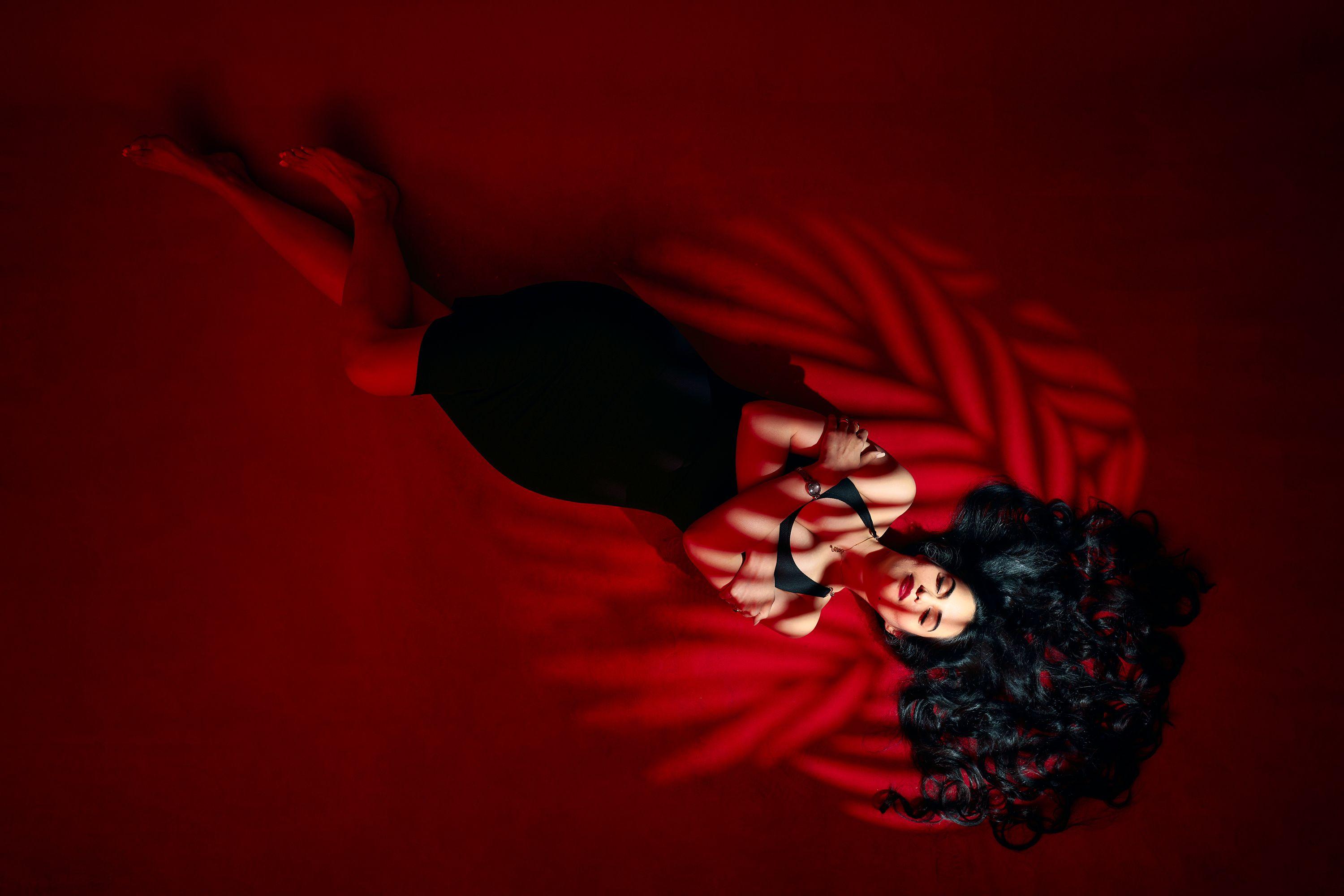 Striking portrait of a woman in a black dress lying on a red background with dramatic shadows.