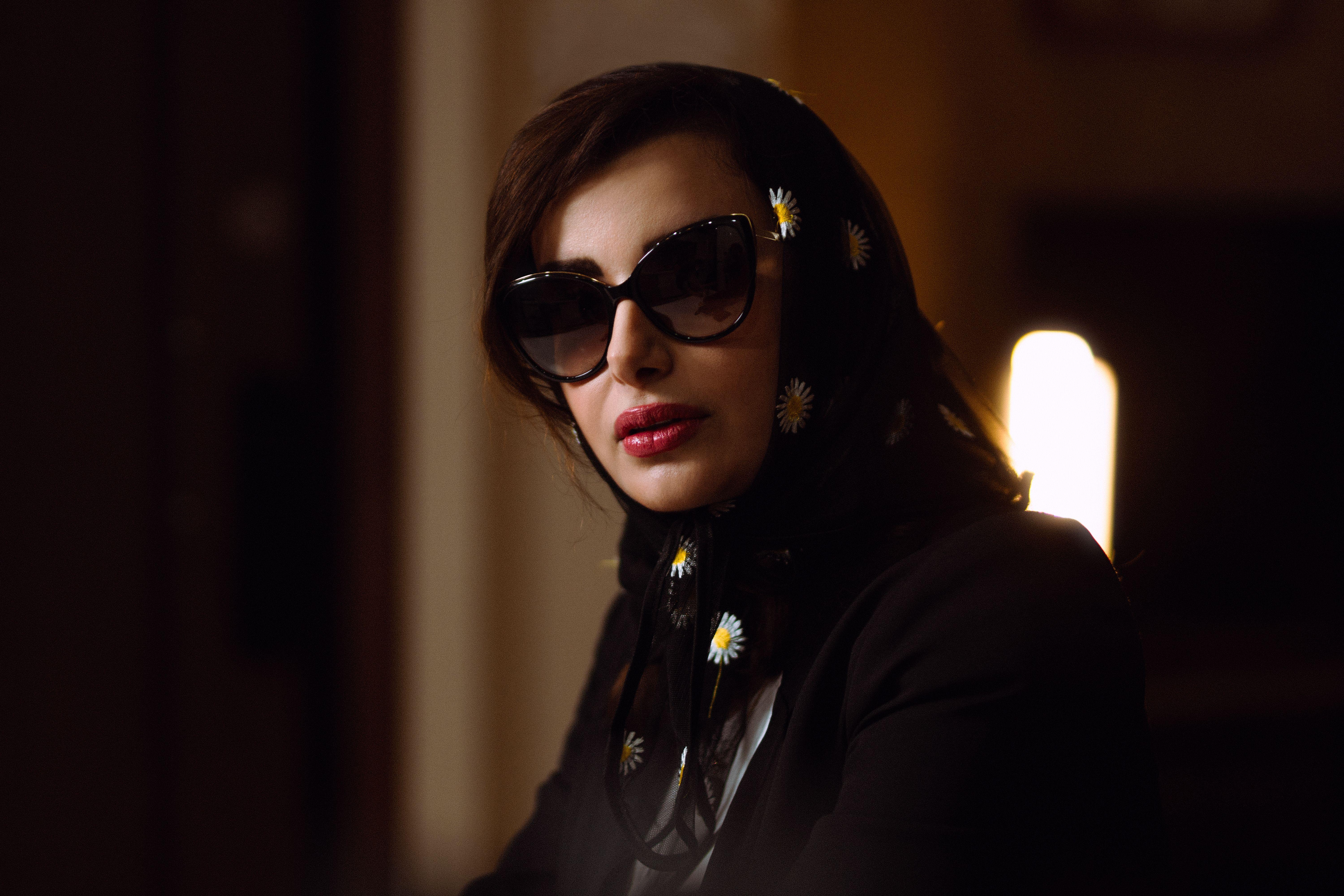 Elegant portrait of a woman wearing sunglasses and a floral shawl in a dimly lit room.