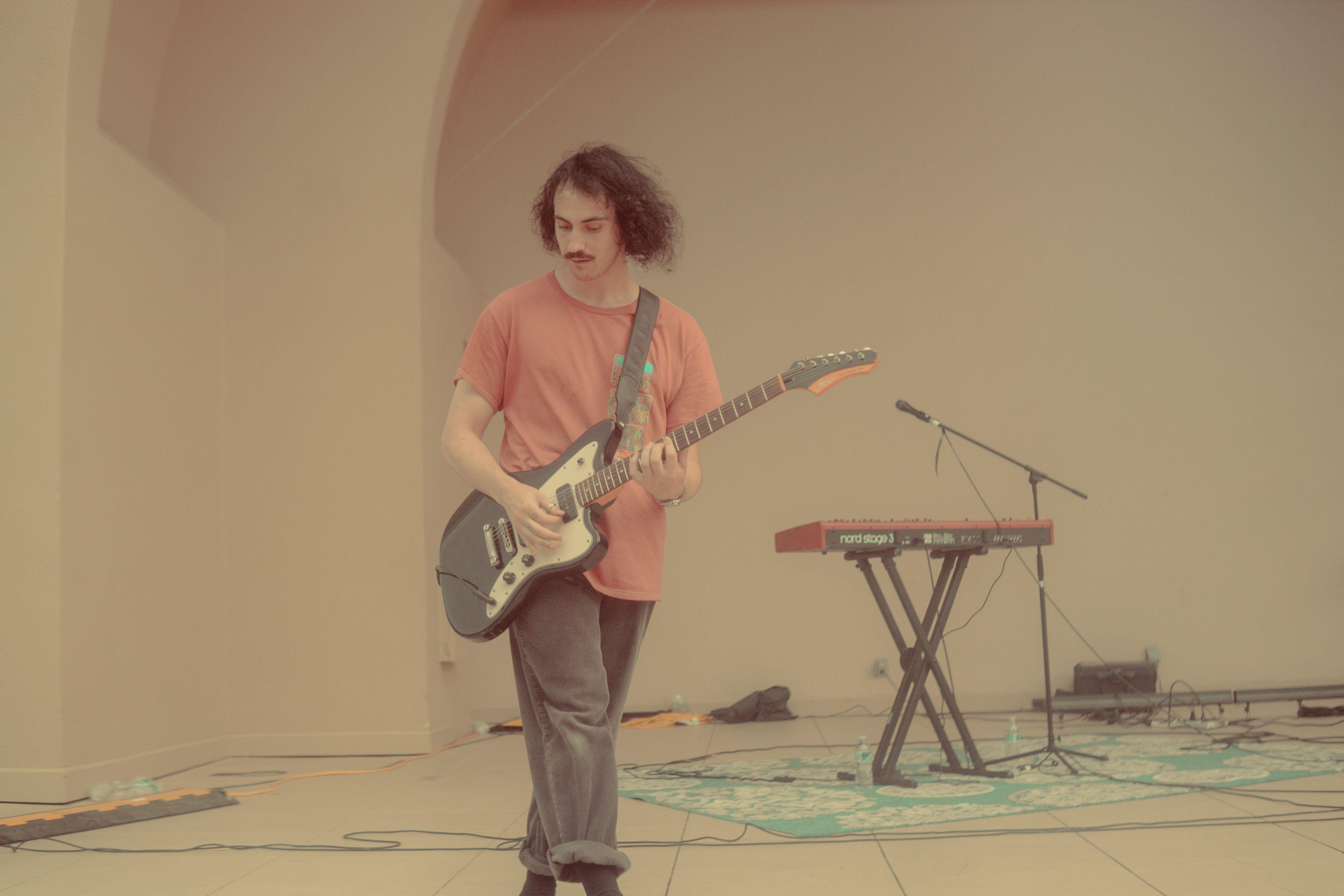 A young musician with a mustache playing an electric guitar on stage indoors.