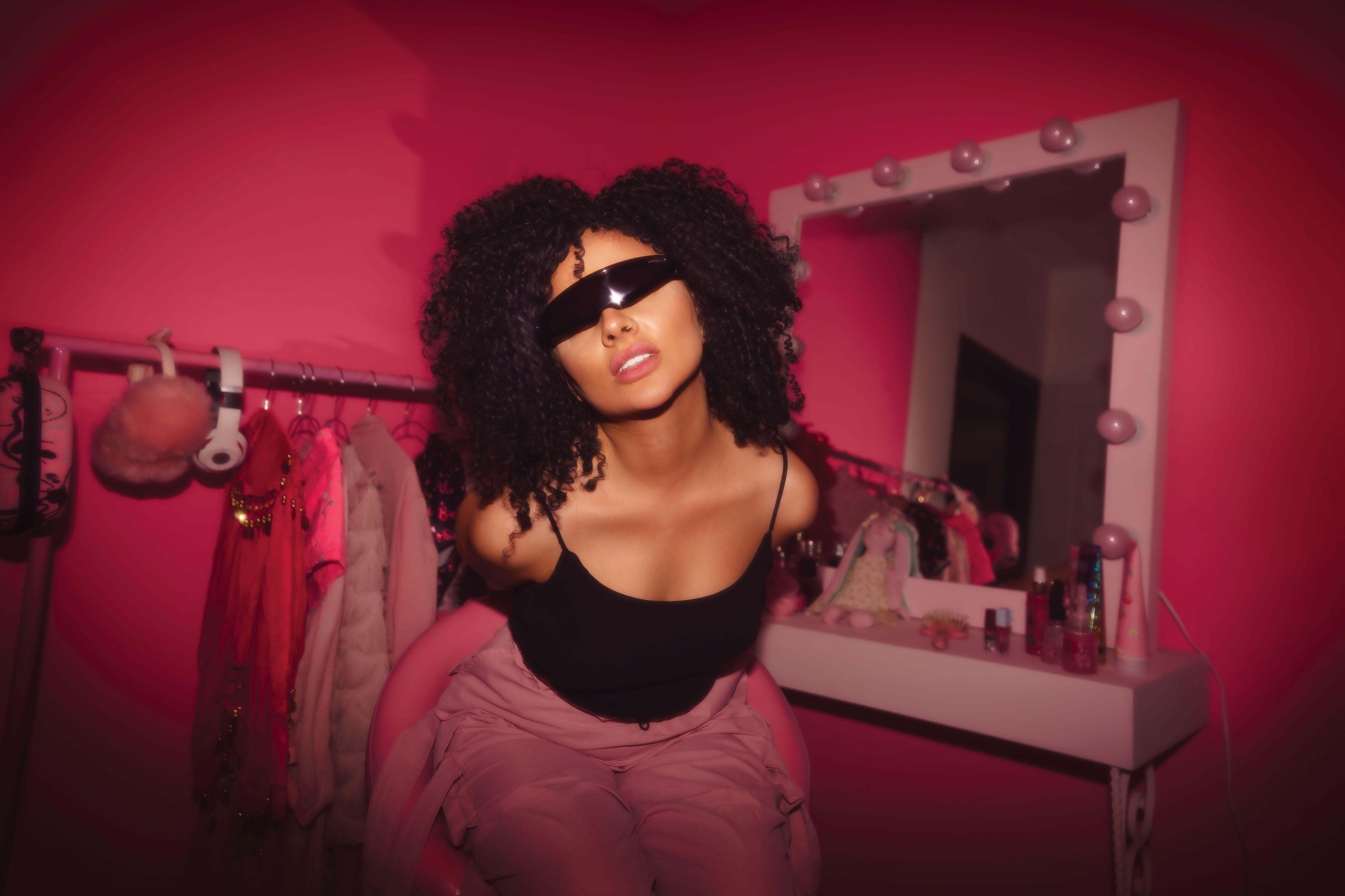 Woman with curly hair and sunglasses posing stylishly in a vibrant pink room.