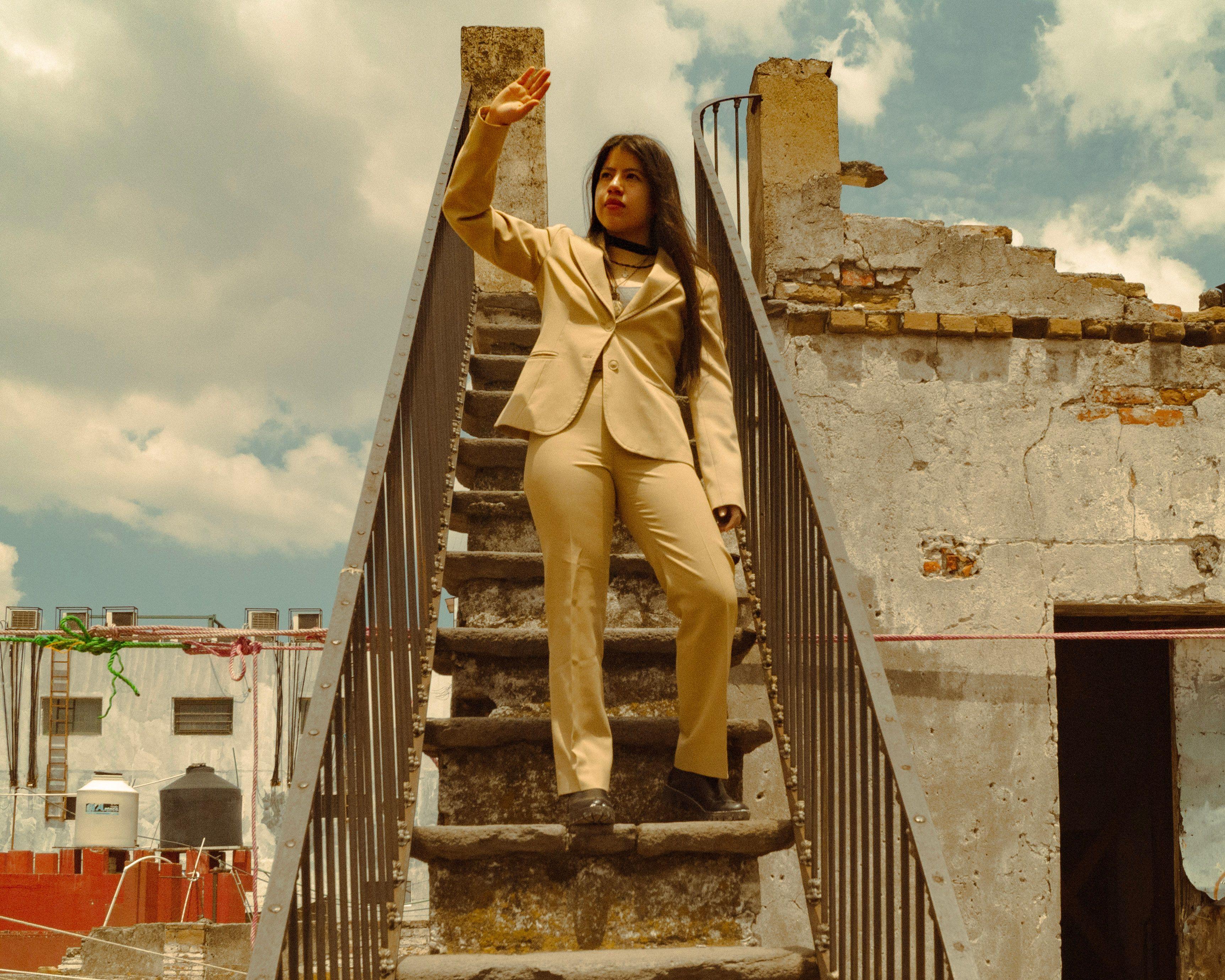 Woman in a beige suit standing on a rooftop staircase against a rustic urban backdrop.