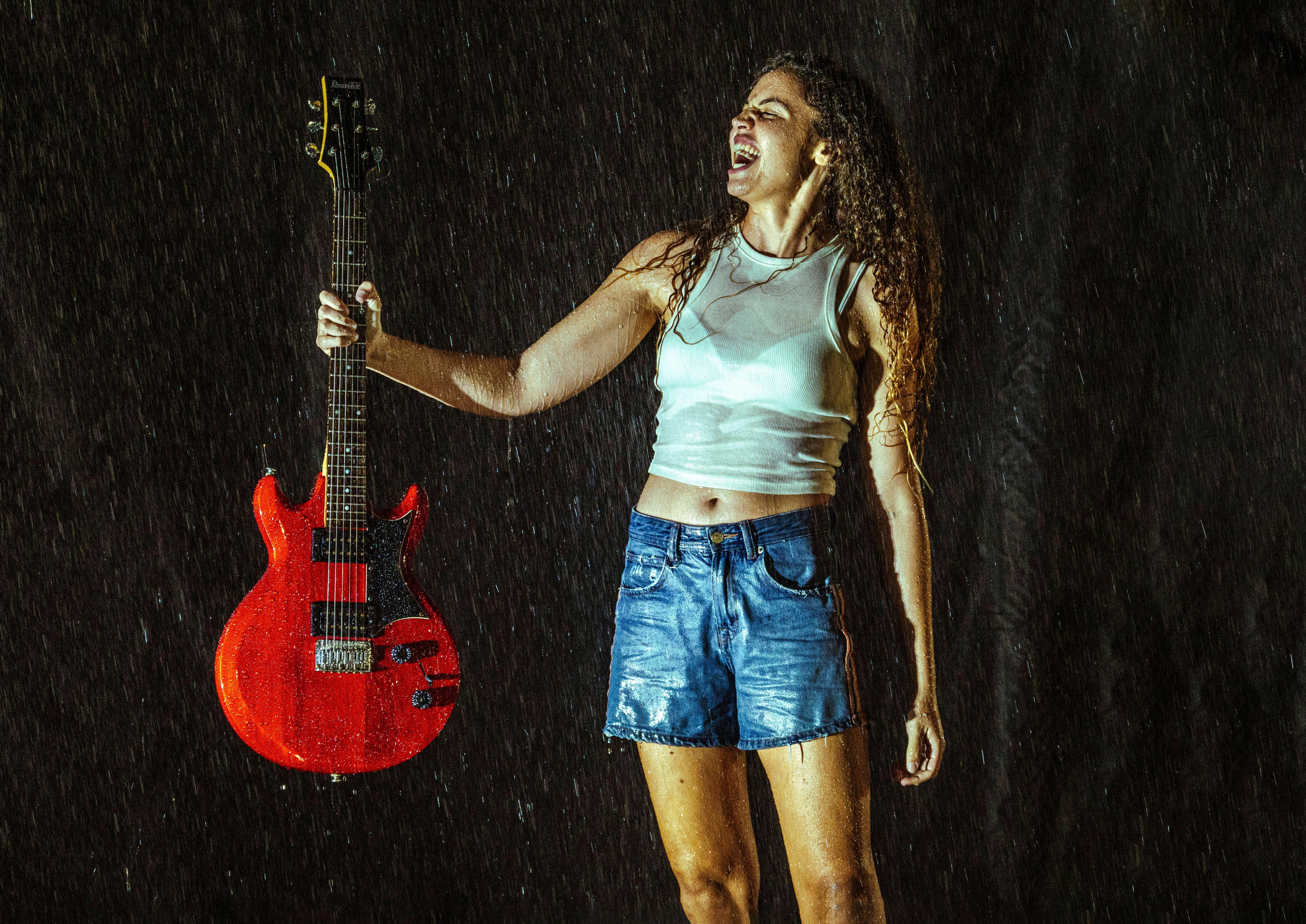 A woman stands boldly in the rain, holding a red electric guitar, under nighttime lighting.
