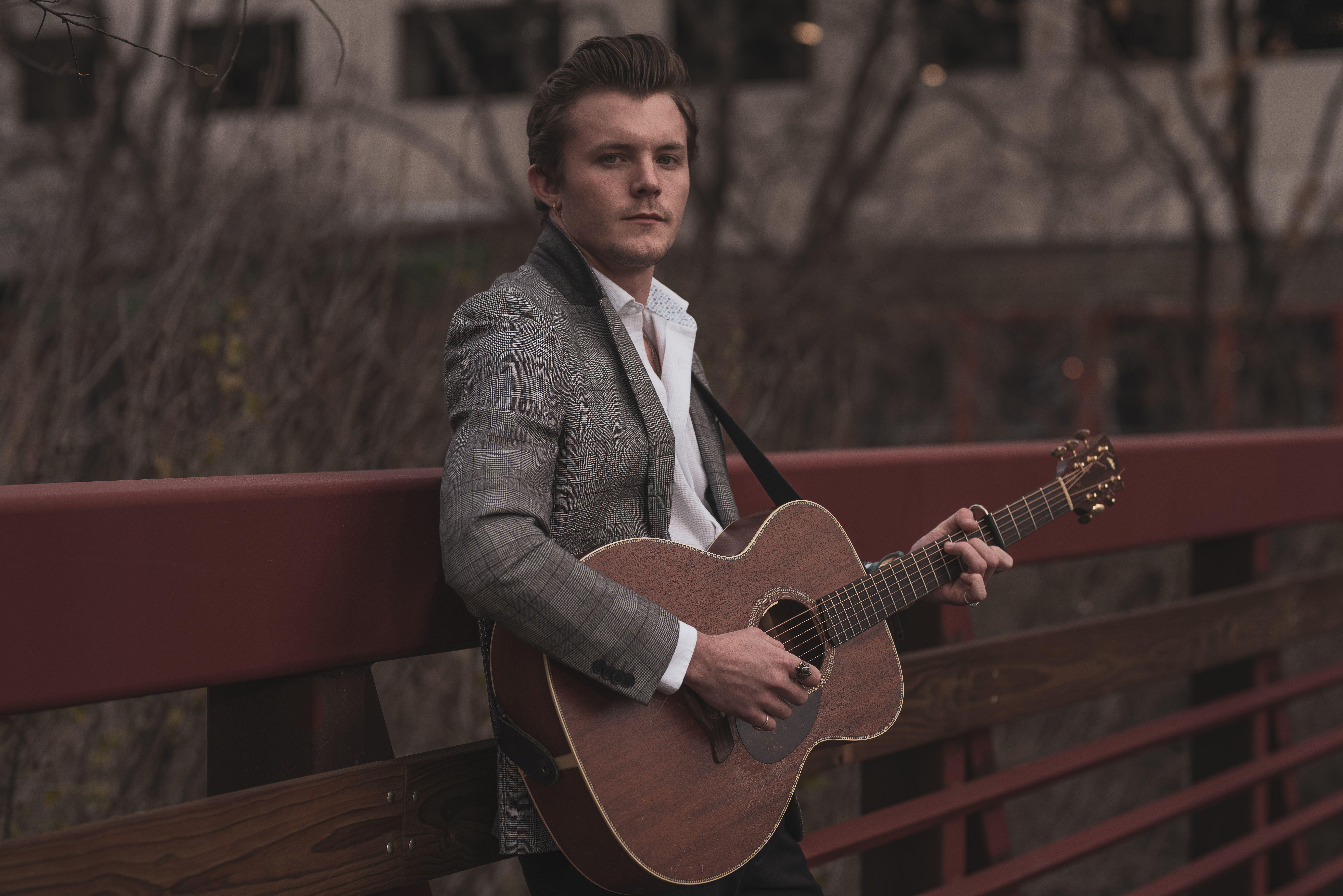 A man playing acoustic guitar on a bridge with a stylish outfit.
