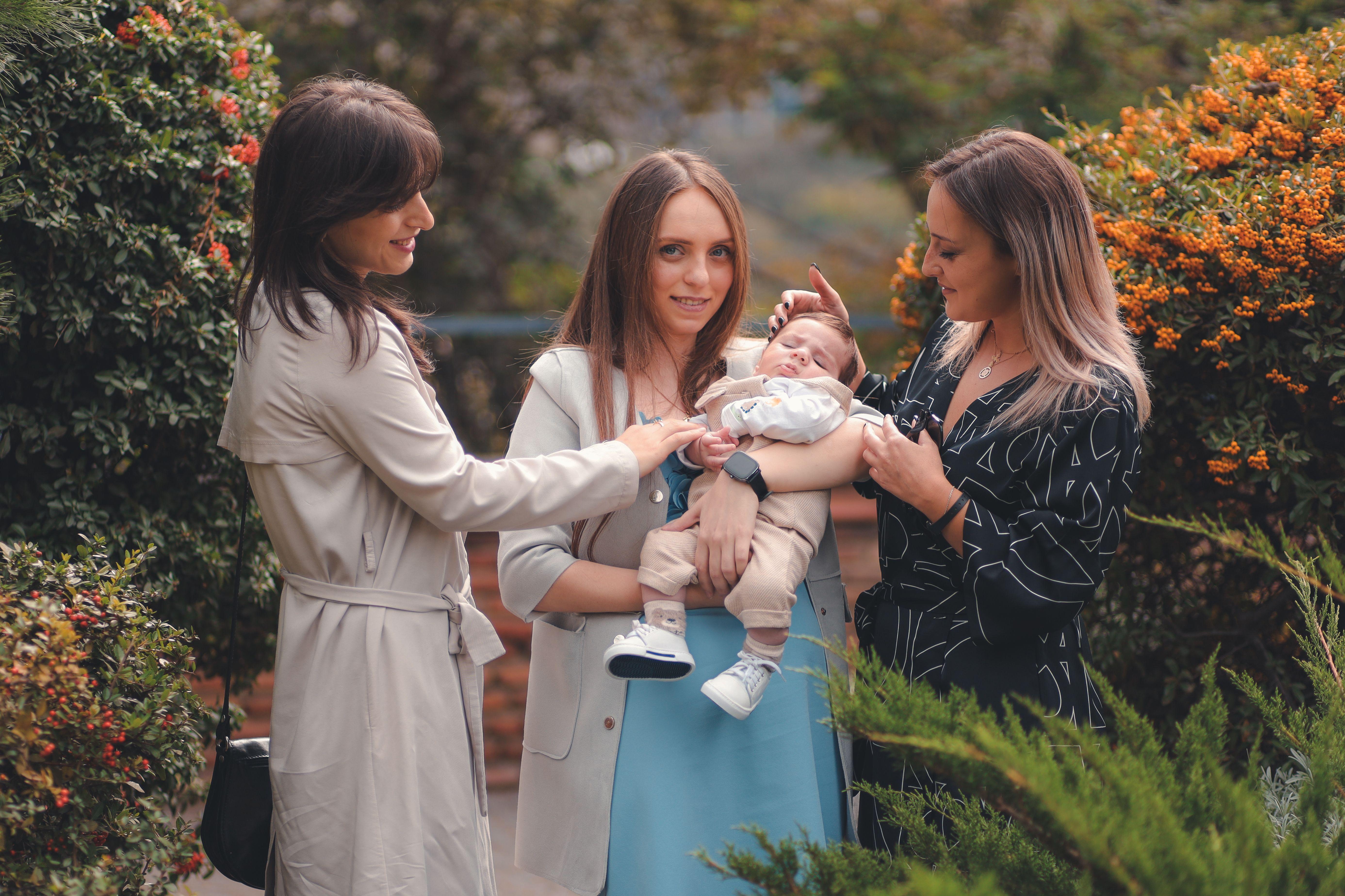 Three women joyfully admiring a newborn in a lush garden. A harmonious blend of nature and family bonding.