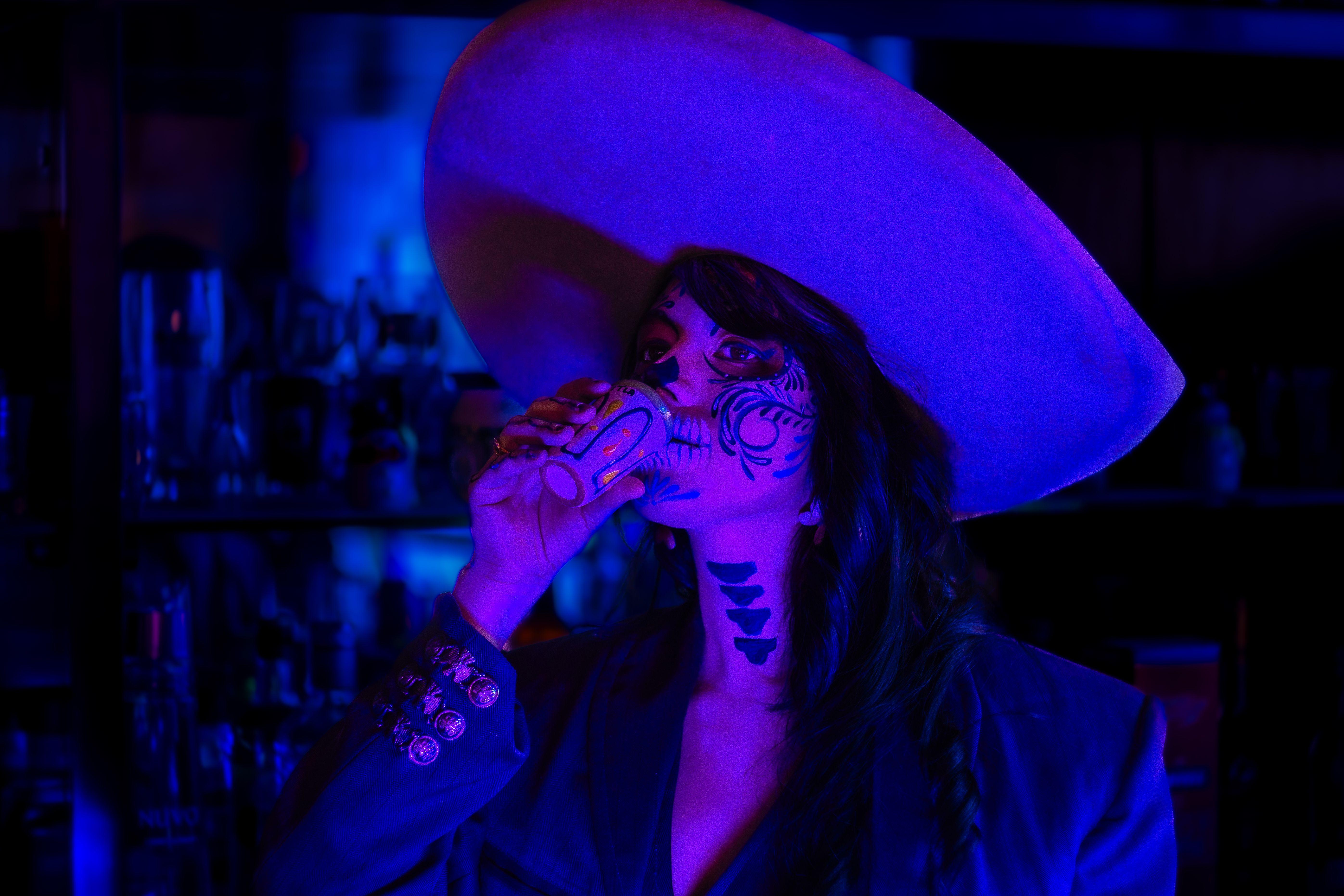 A woman with elegant Catrina makeup drinks in neon blue light, celebrating Mexican culture.