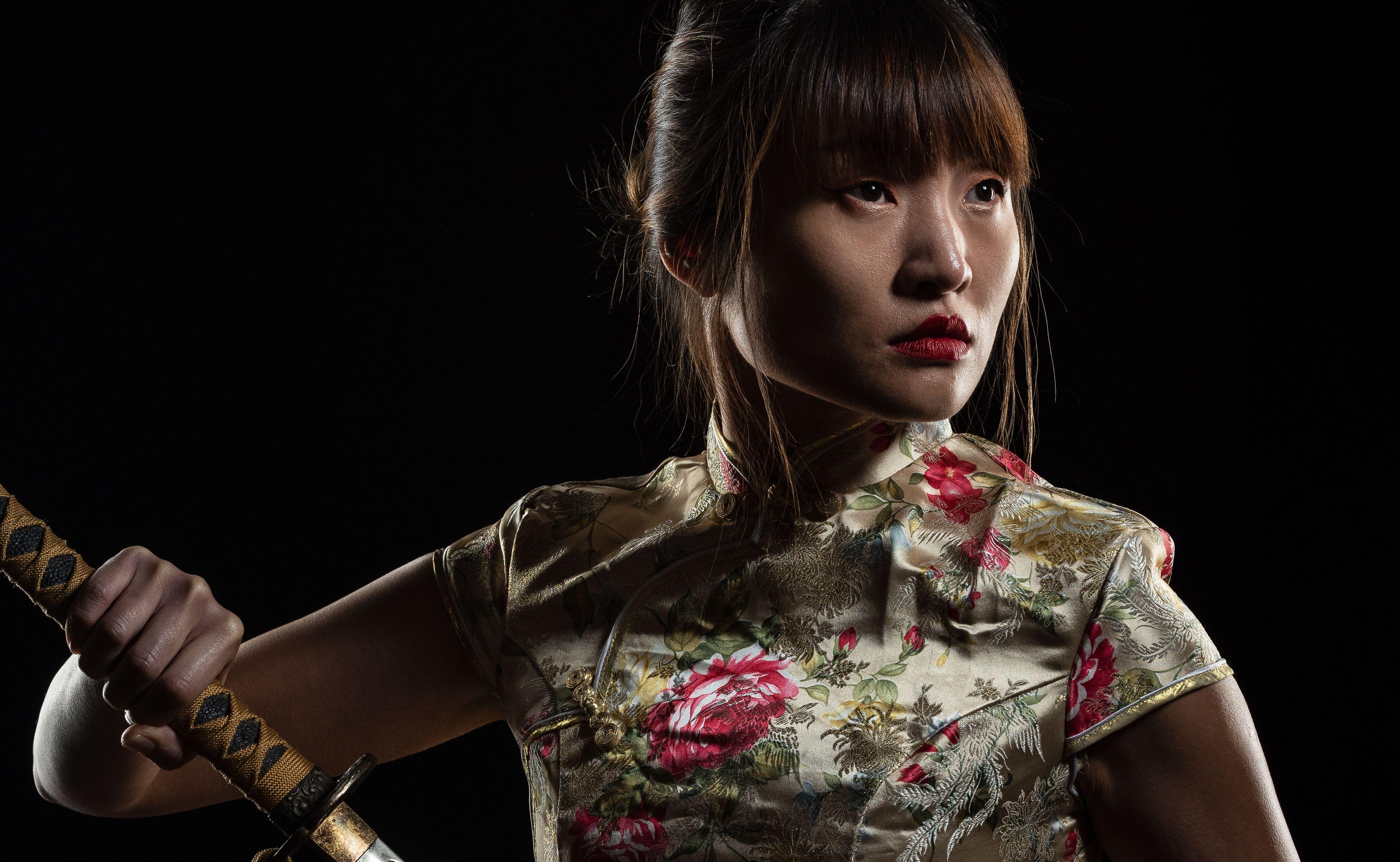 Portrait of a Japanese woman in traditional clothing, holding a katana, captured dramatically against a black background.