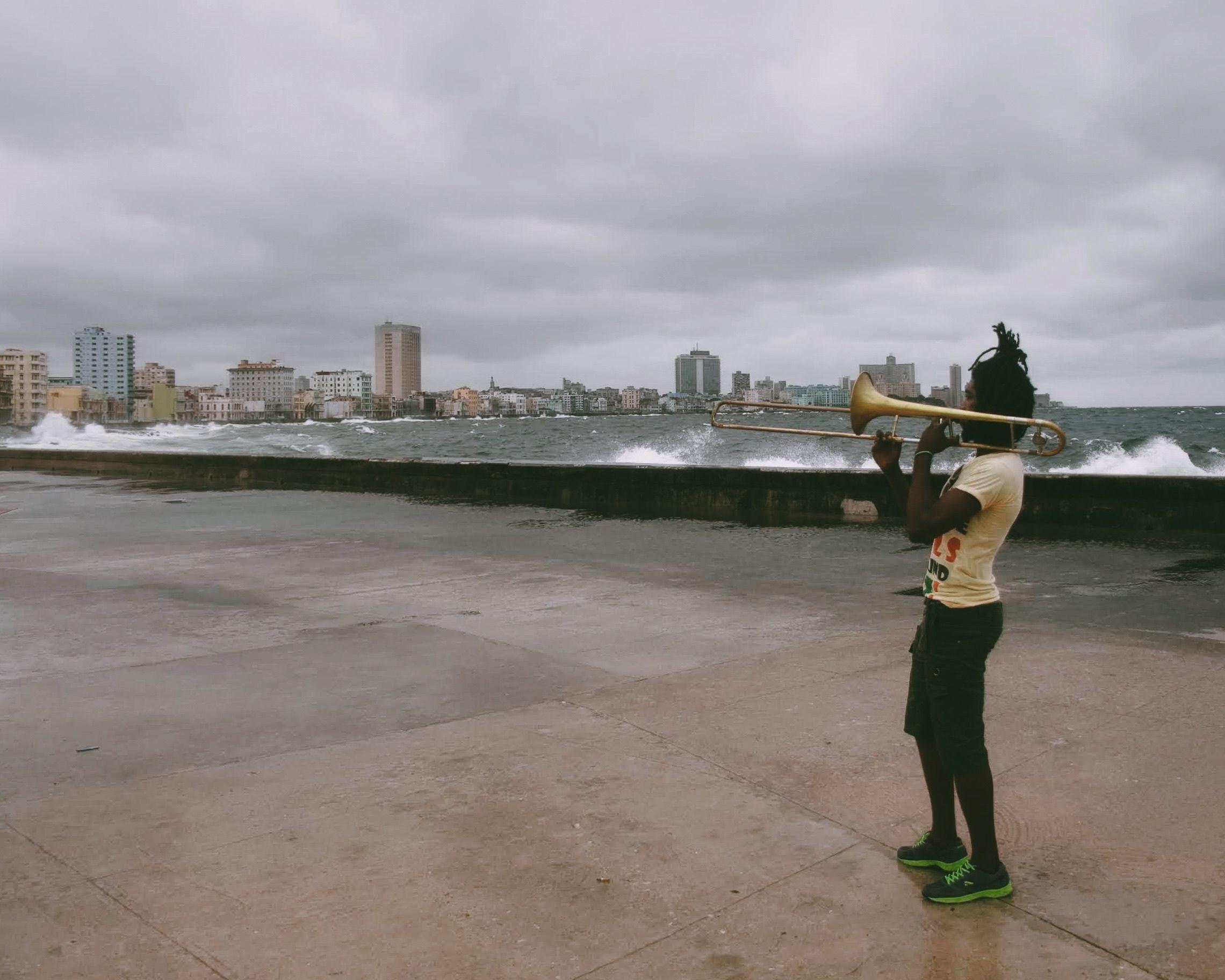 Young man playing trombone outdoors near the ocean on a cloudy day.
