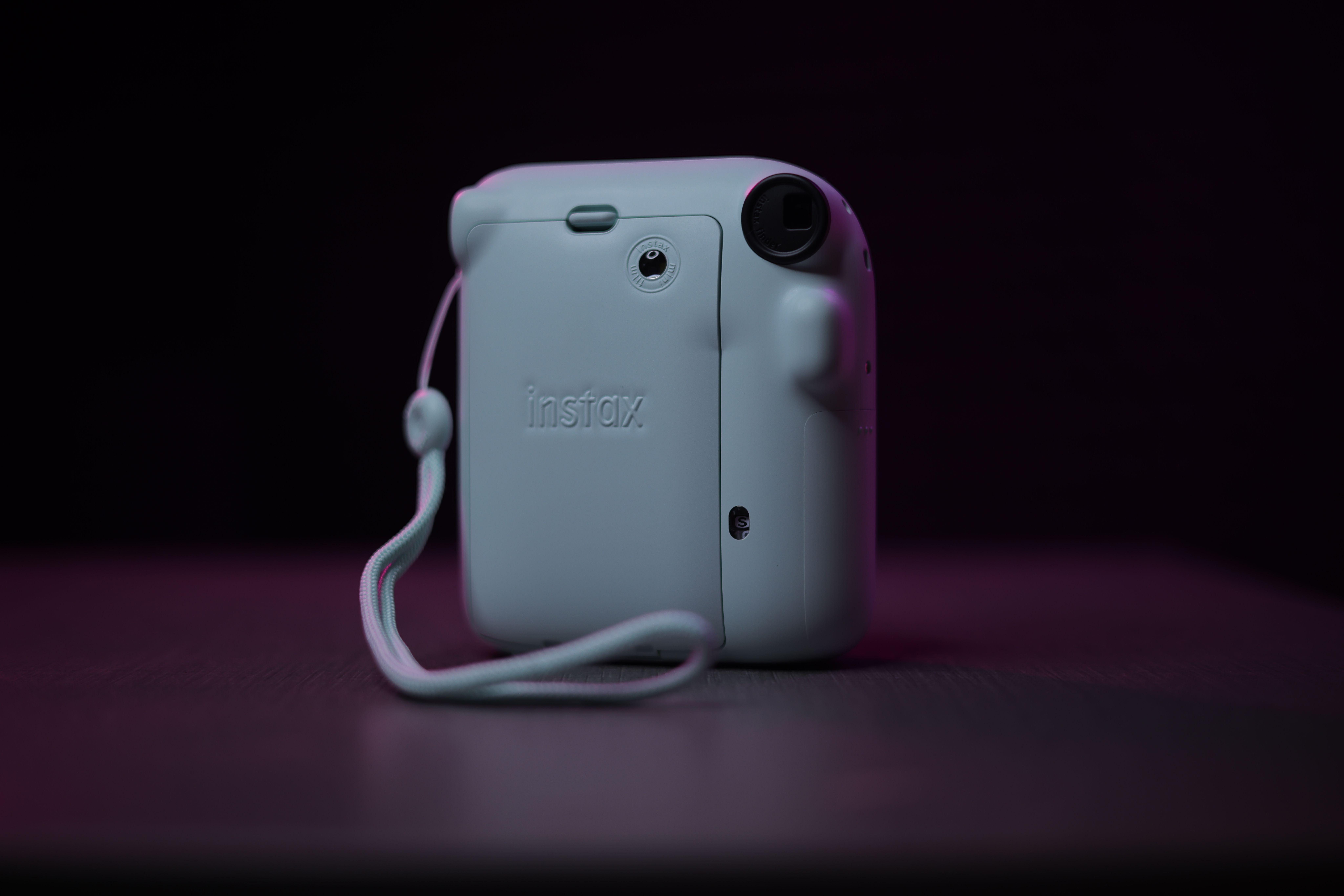 A close-up shot of a Fujifilm Instax Mini camera with a wrist strap, set against a dark background.