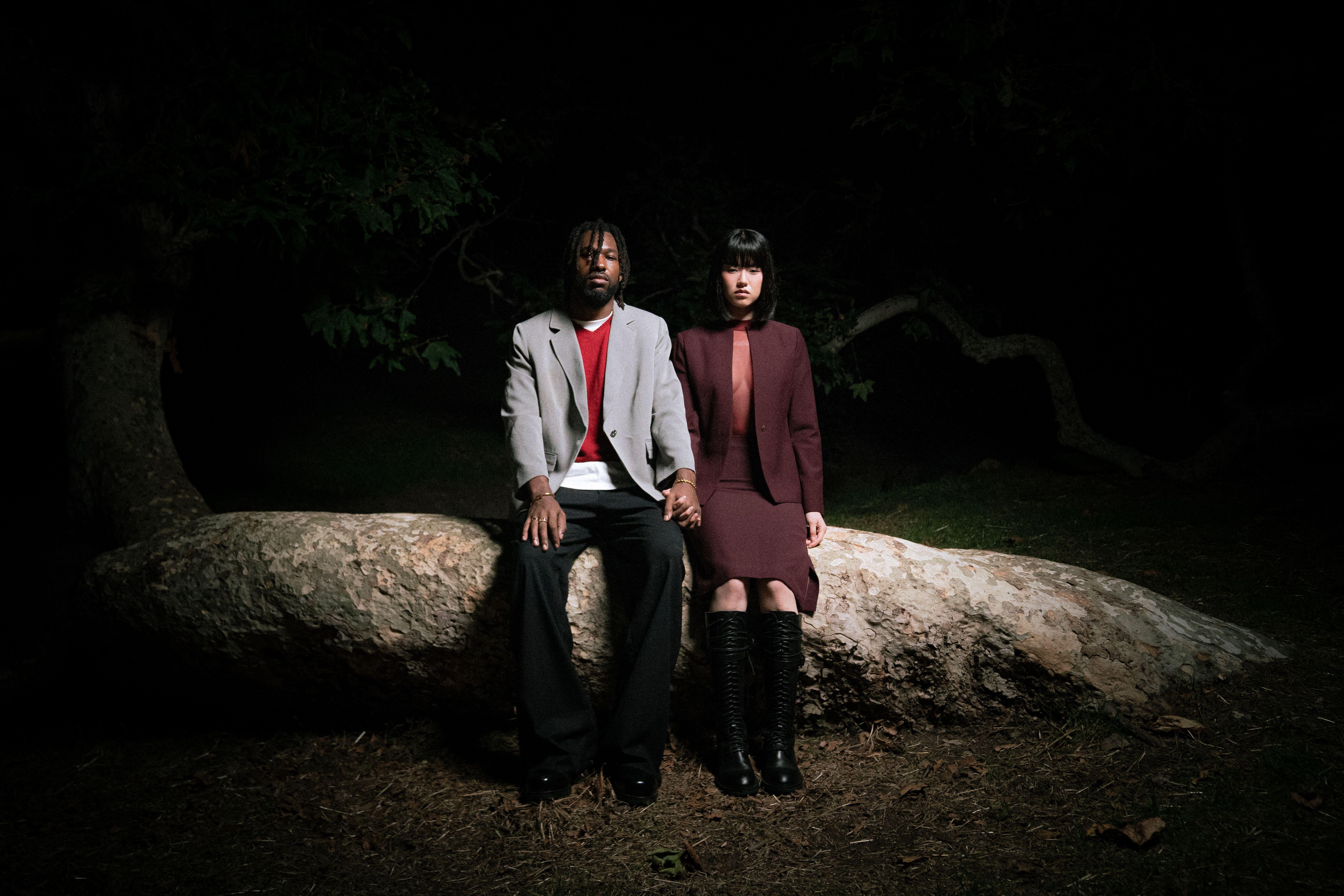 A stylish couple in formal attire seated on a large tree trunk under night skies, holding hands.