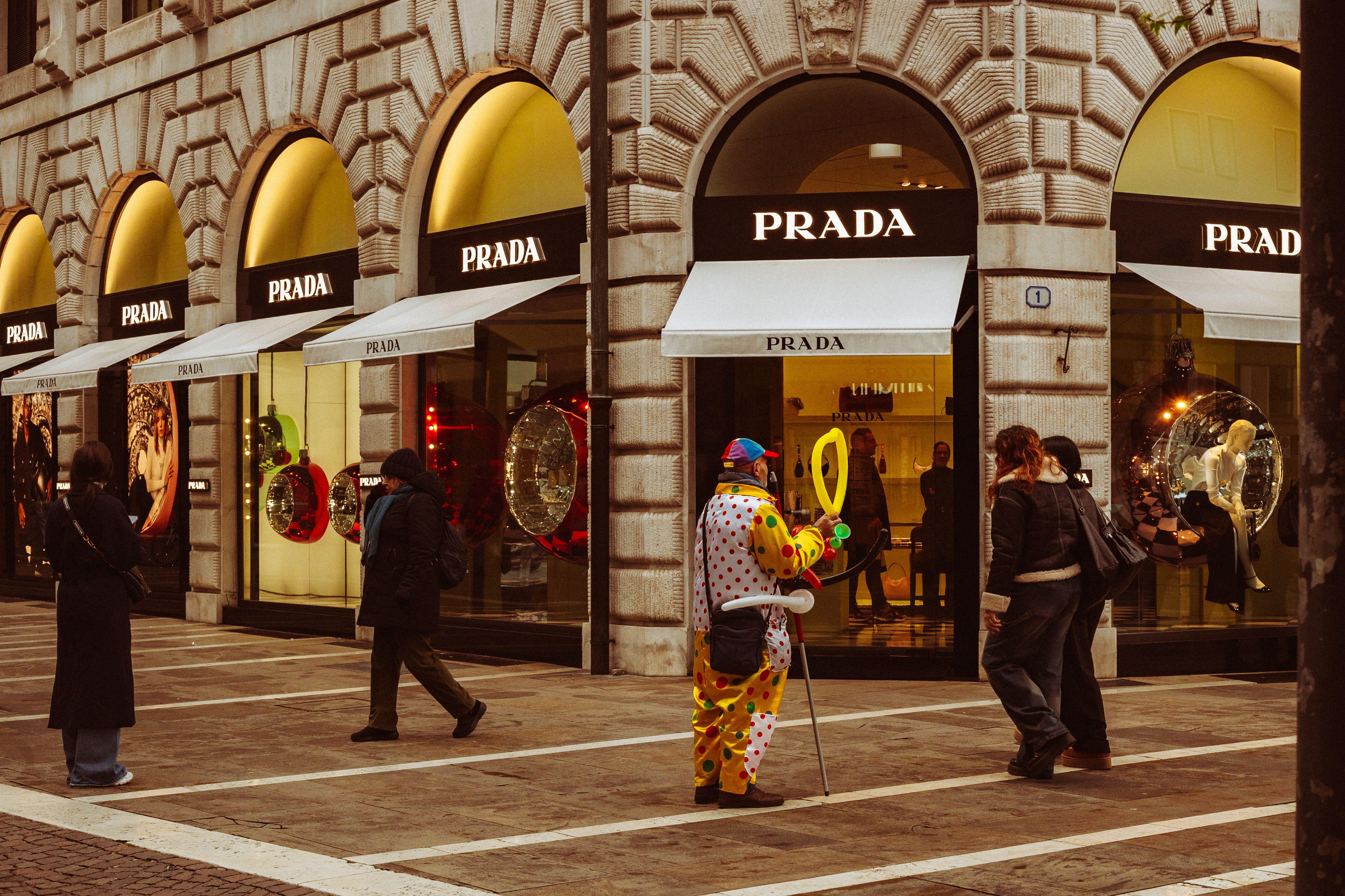 People passing by a vibrant clown entertainer outside a Prada store in Padova, Italy.