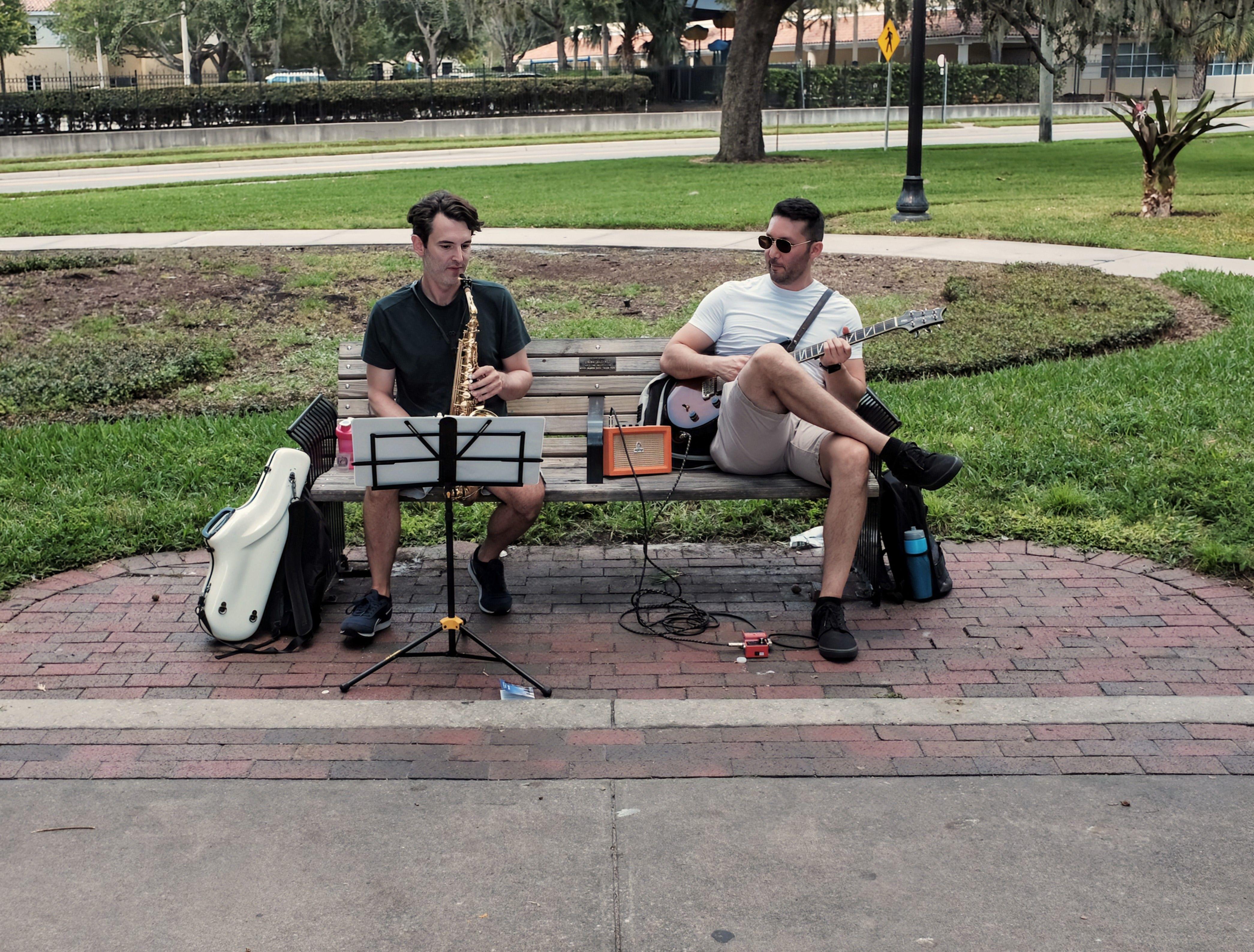 Two musicians playing guitar and saxophone on a park bench in Orlando, FL.
