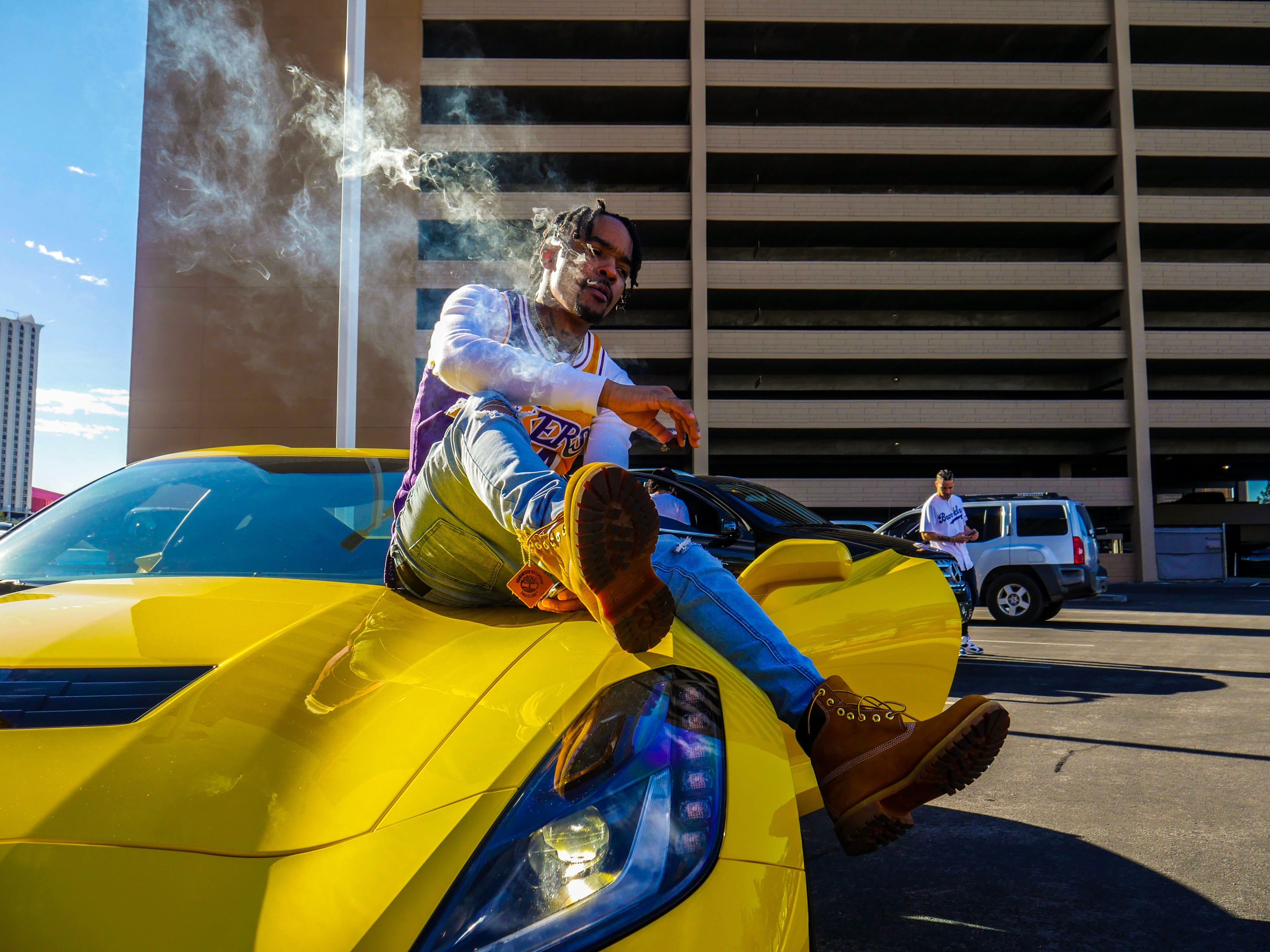 A fashionable man sits on a yellow sportscar in an urban setting, exuding style and confidence.