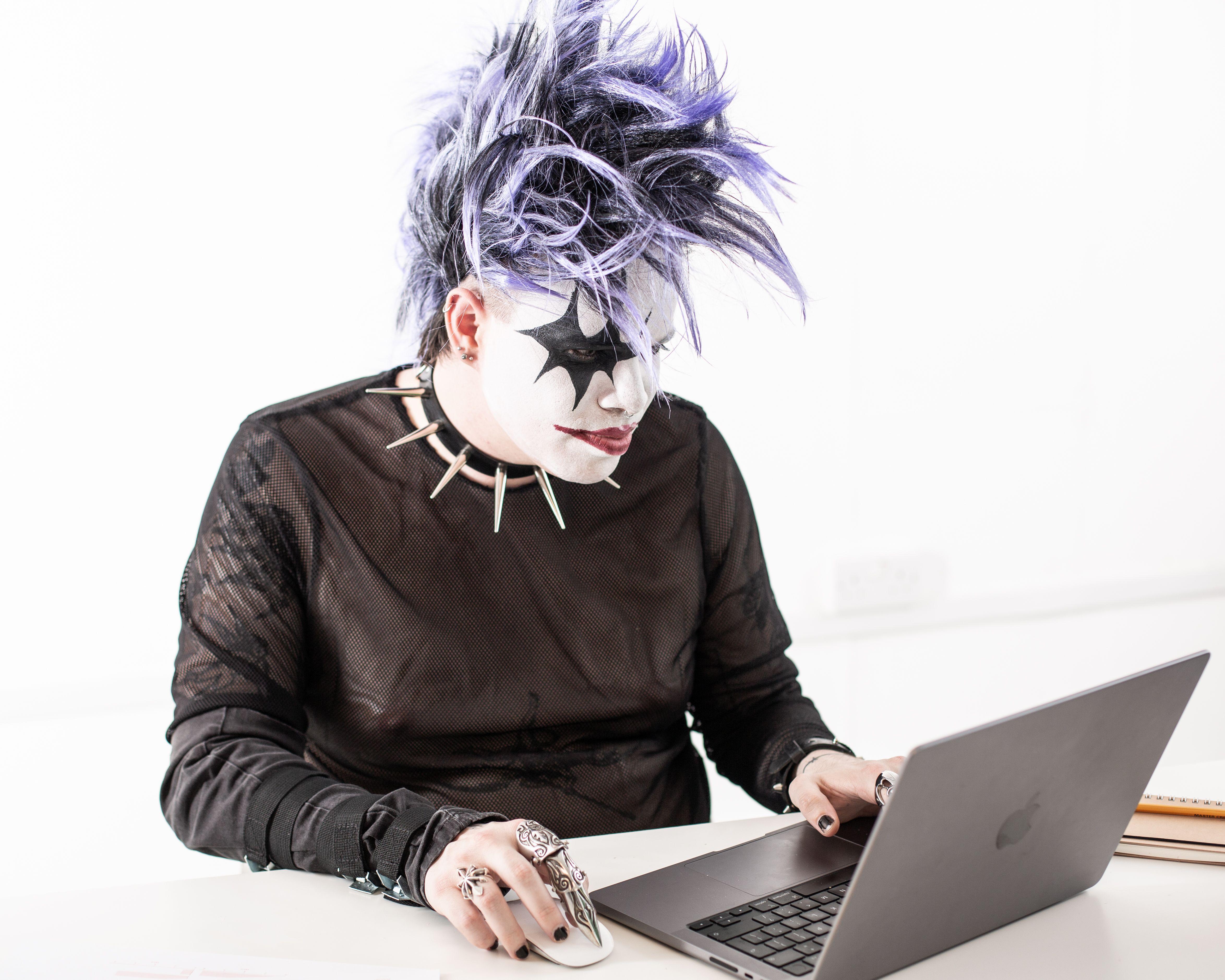 A person with costume makeup and purple hair working on a laptop against a white background.