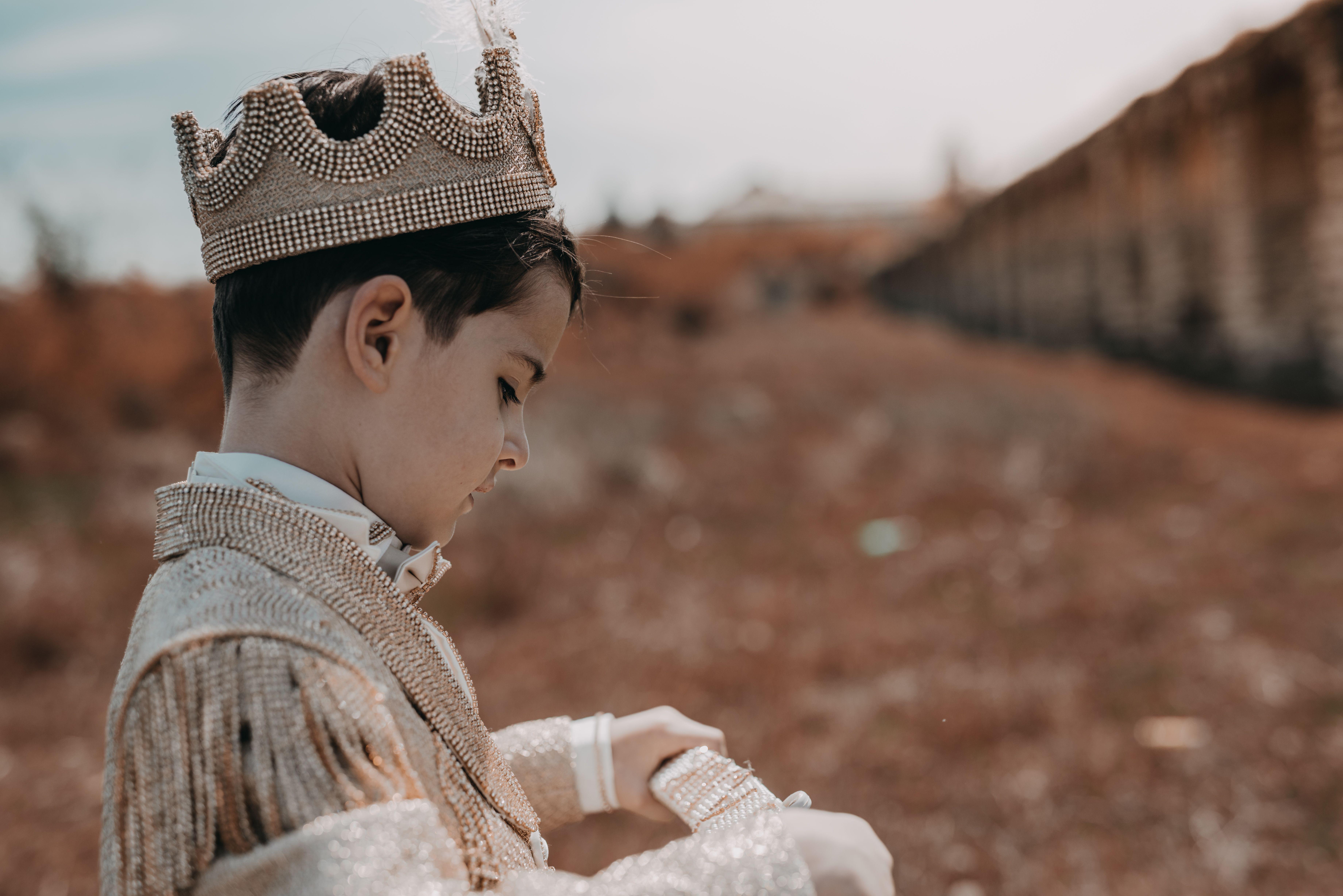 A child dressed as a prince wearing a crown in an outdoor setting with fall scenery.