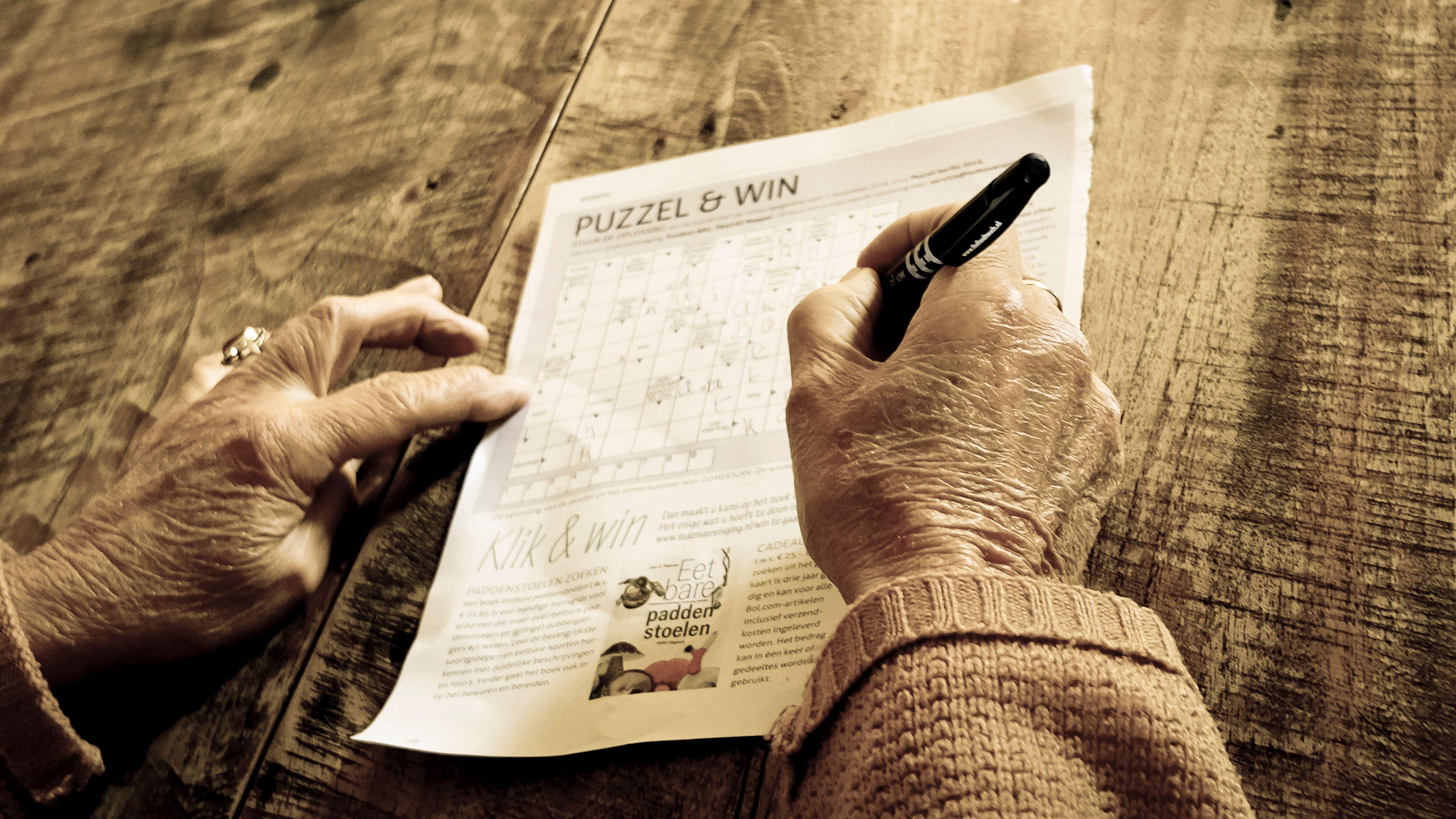 Close-up of a senior adult solving a puzzle on a rustic wooden table with a pen.